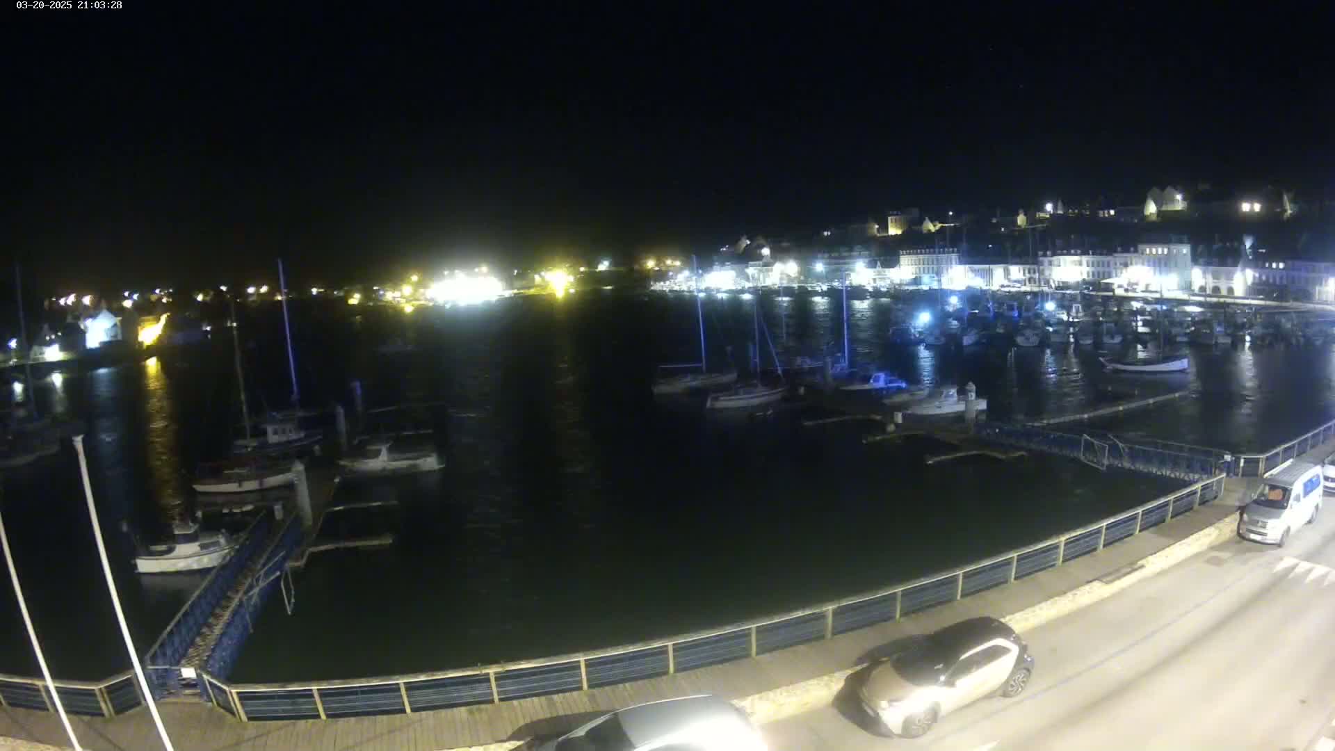 A nighttime view of a harbor filled with boats, a town with lights illuminating the buildings in the background, and cars parked along a roadway next to the water.
