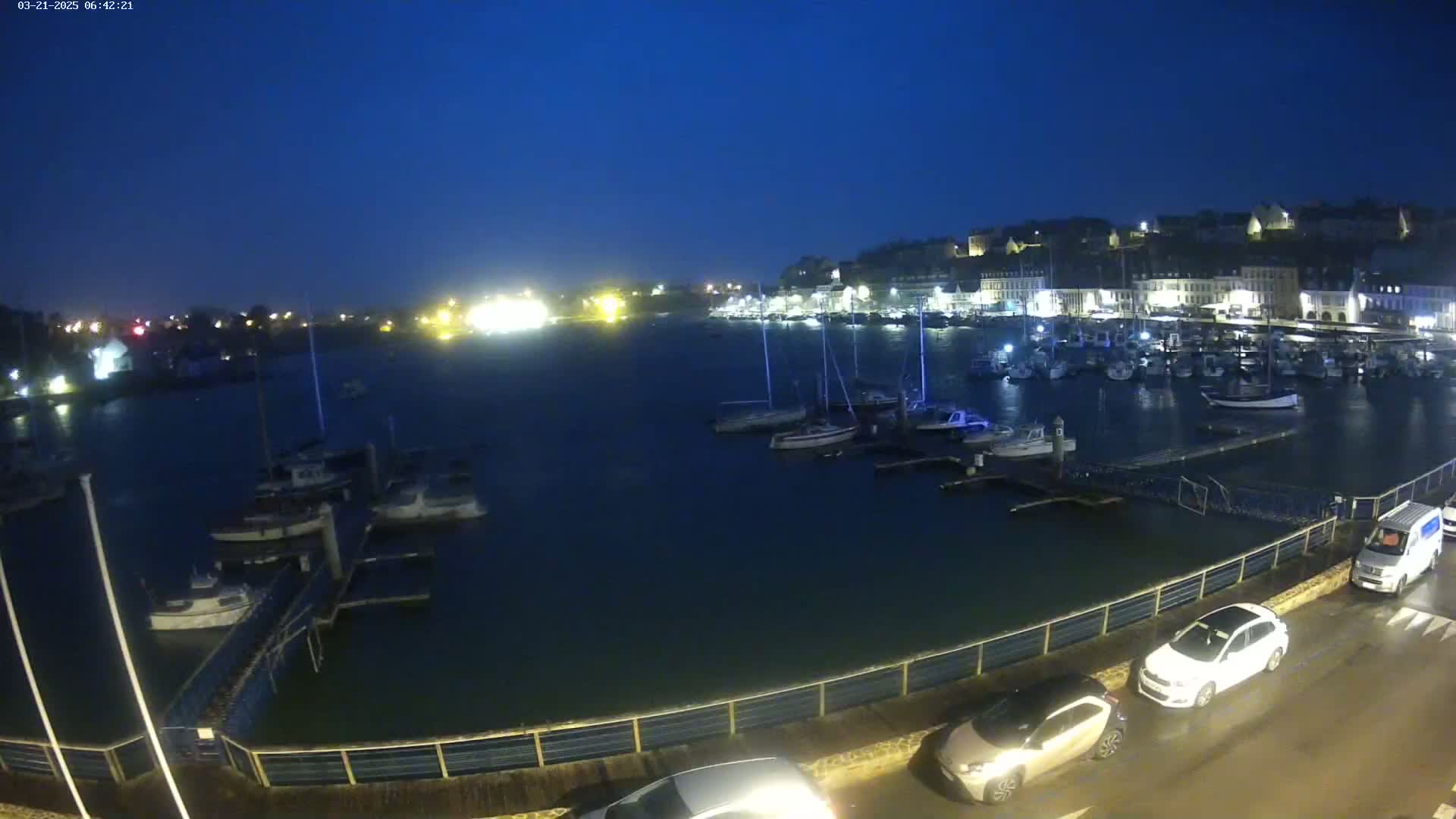A harbor filled with boats is seen at night under a dark blue sky, with cars parked along a nearby roadway.
