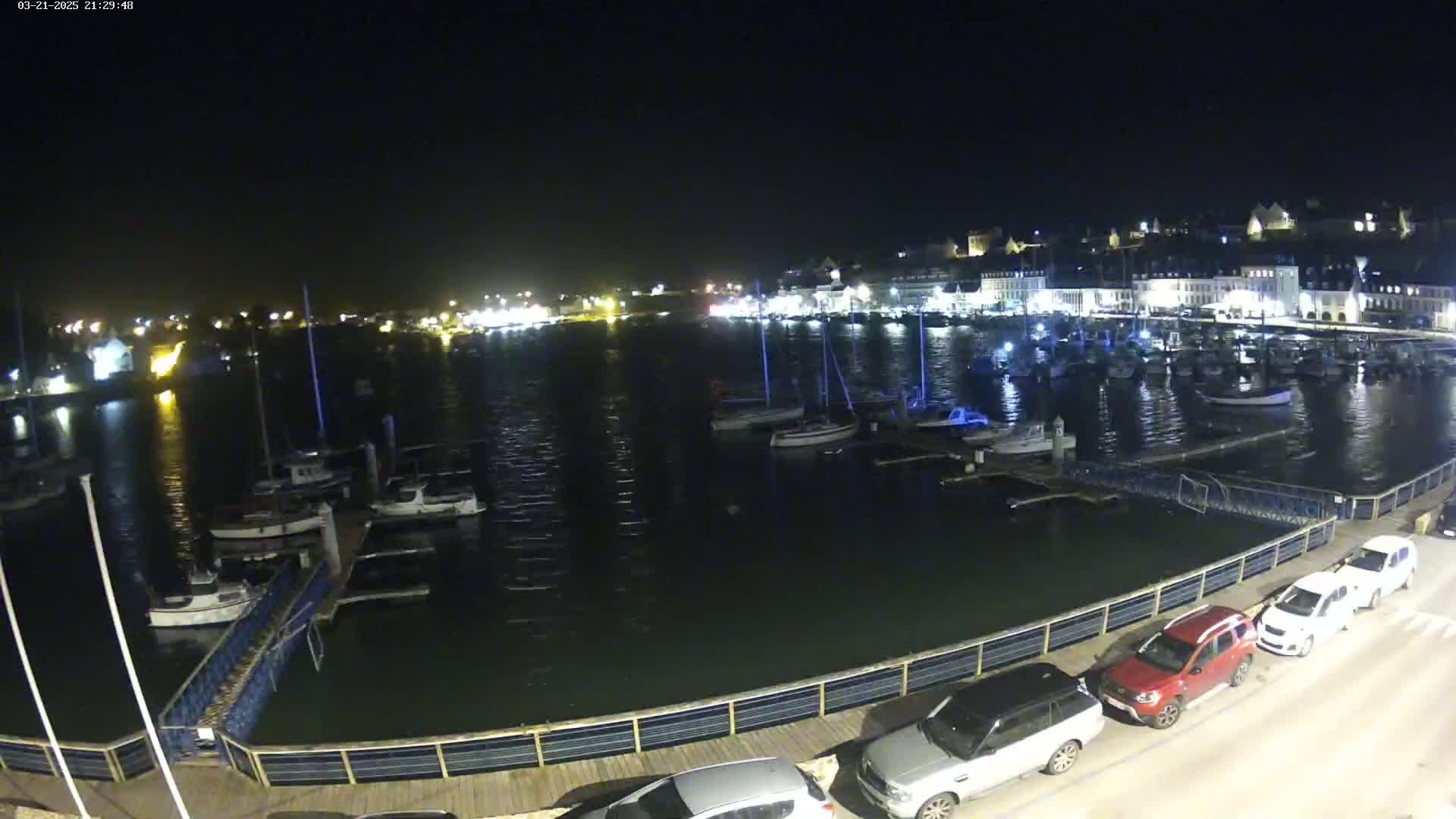 A nighttime view of a harbor filled with boats, a town with lights, and several cars parked along a waterfront boardwalk under a clear, dark sky.