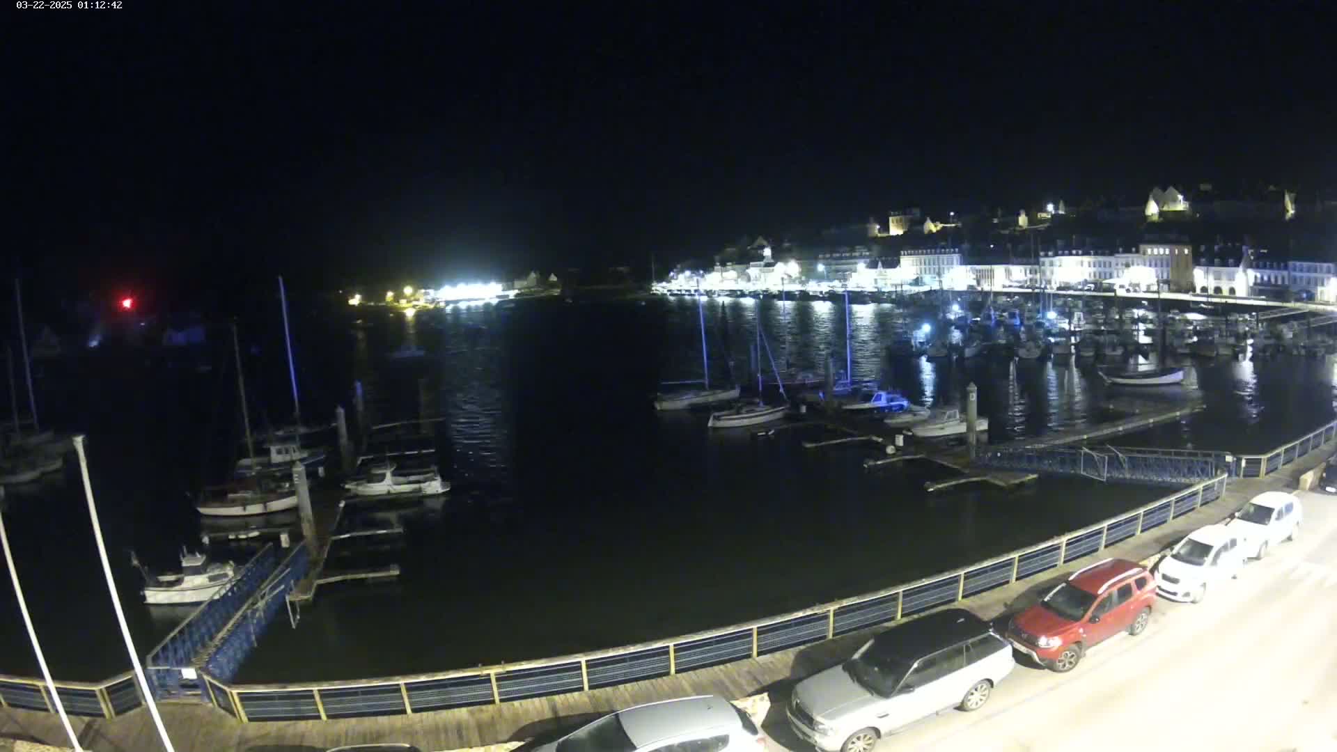 A nighttime view of a harbor filled with boats, docked along a waterfront with cars parked nearby under a clear sky.