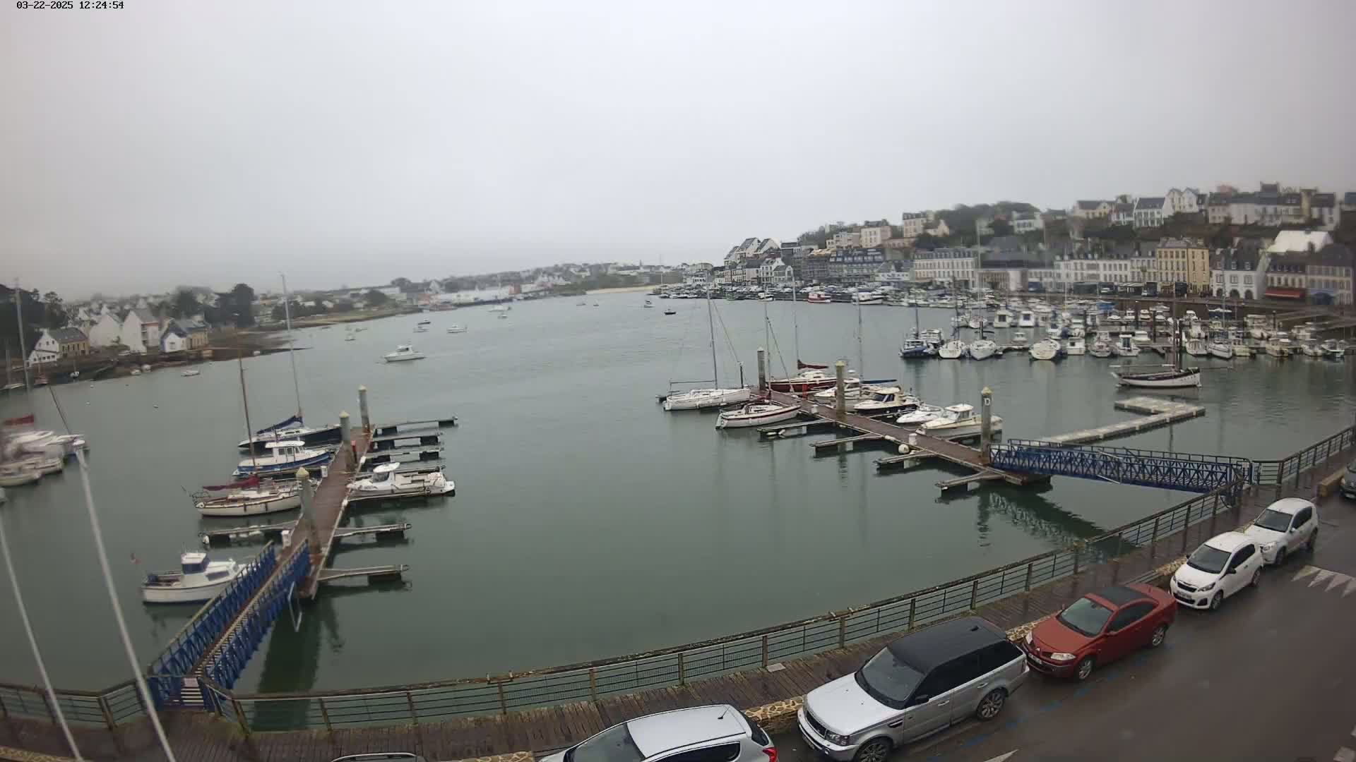 A harbor filled with boats on a cloudy day, with cars parked along a nearby walkway overlooking the water.