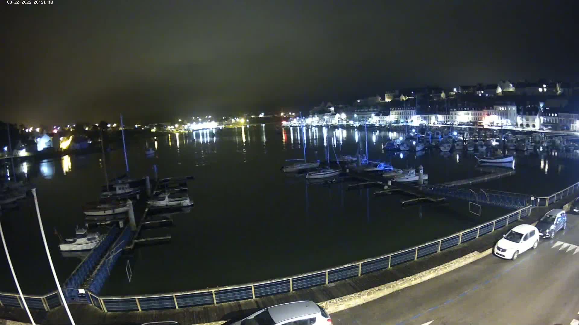 A nighttime view of a harbor filled with boats, lit by city lights across the water under a dark, cloudy sky.