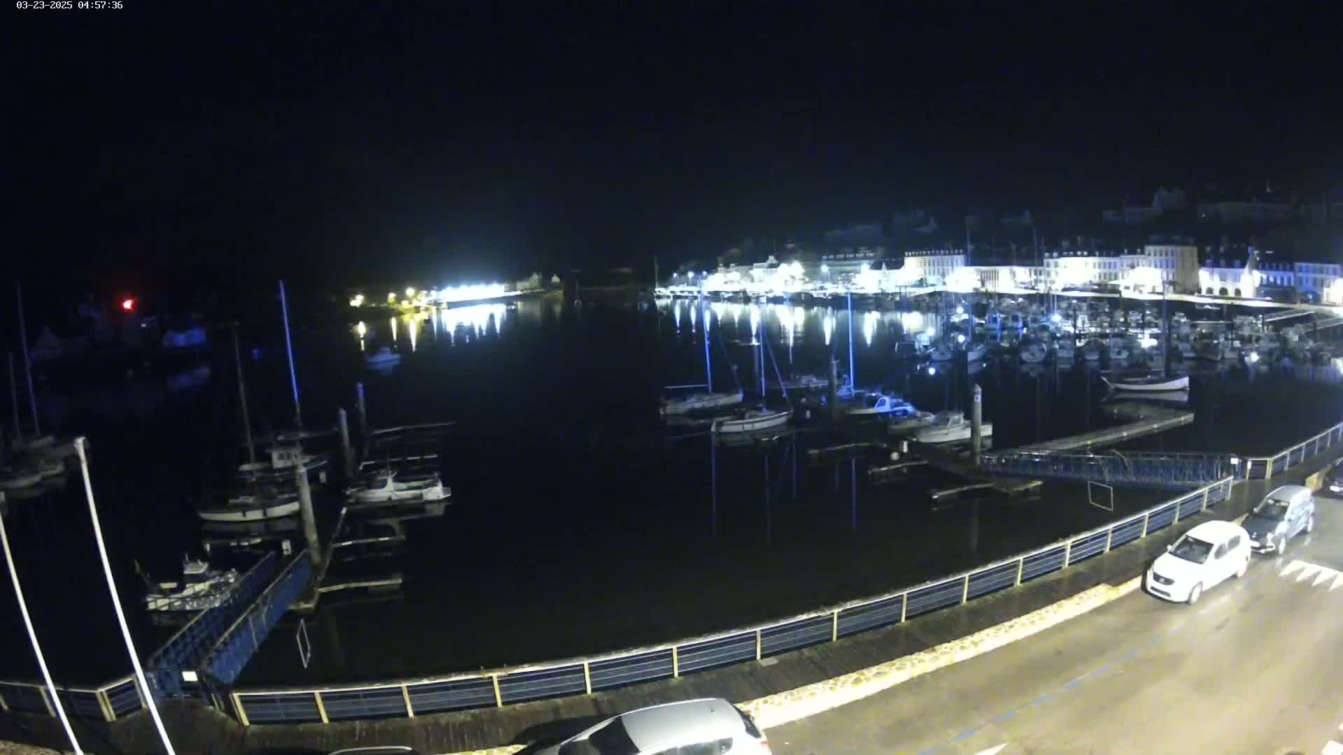 A nighttime view of a harbor filled with boats, docked along a waterfront illuminated by lights, with a few cars parked nearby on a dark, wet road.