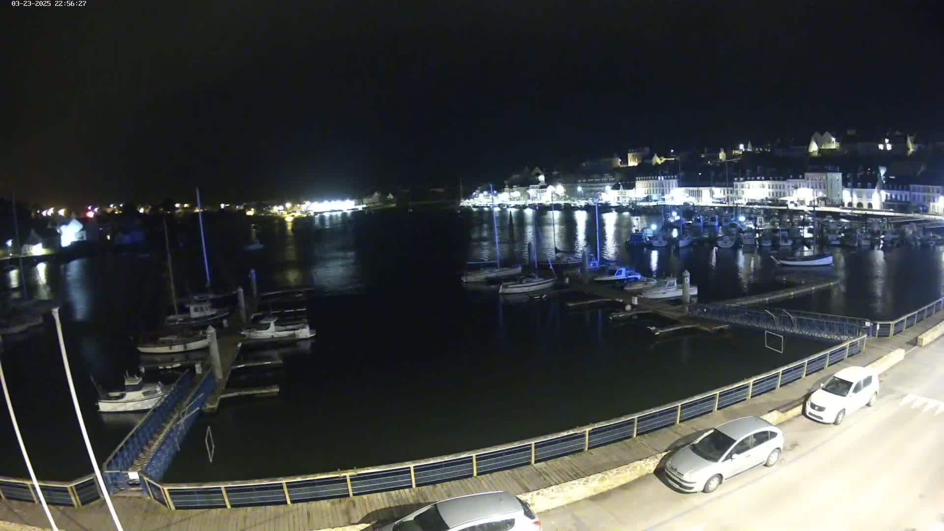 A nighttime view of a harbor filled with boats, docked along a waterfront with a few cars parked nearby under a clear, dark sky.