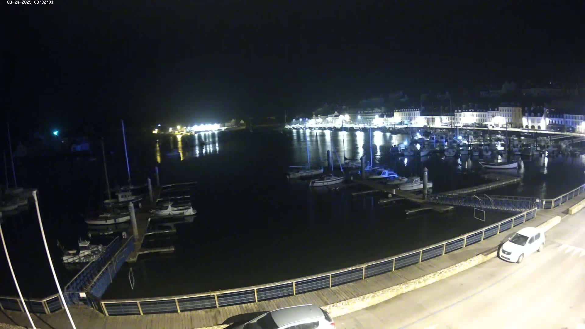A nighttime view of a harbor filled with boats docked along a waterfront with buildings and streetlights visible in the background under a clear, dark sky.