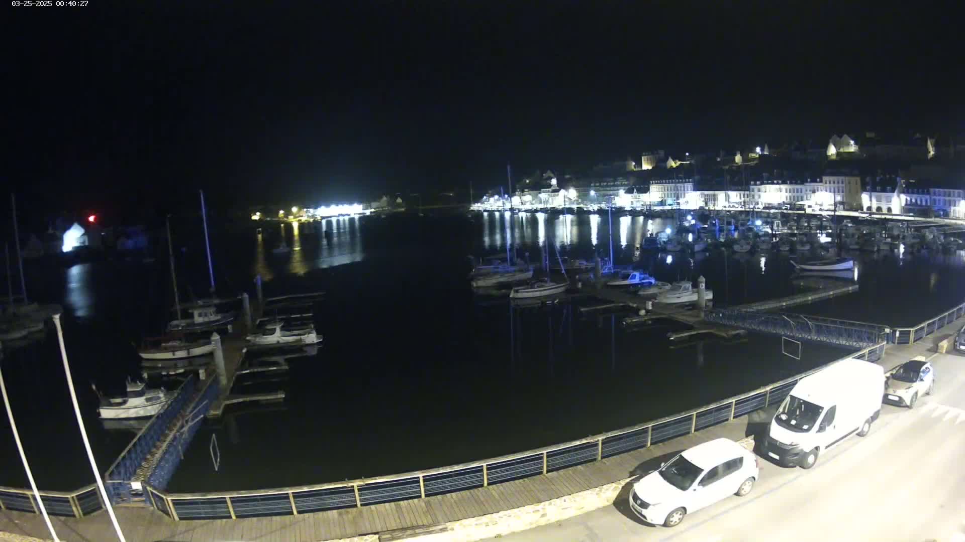 A nighttime view of a harbor filled with boats, alongside a waterfront with parked cars and a town illuminated in the background.
