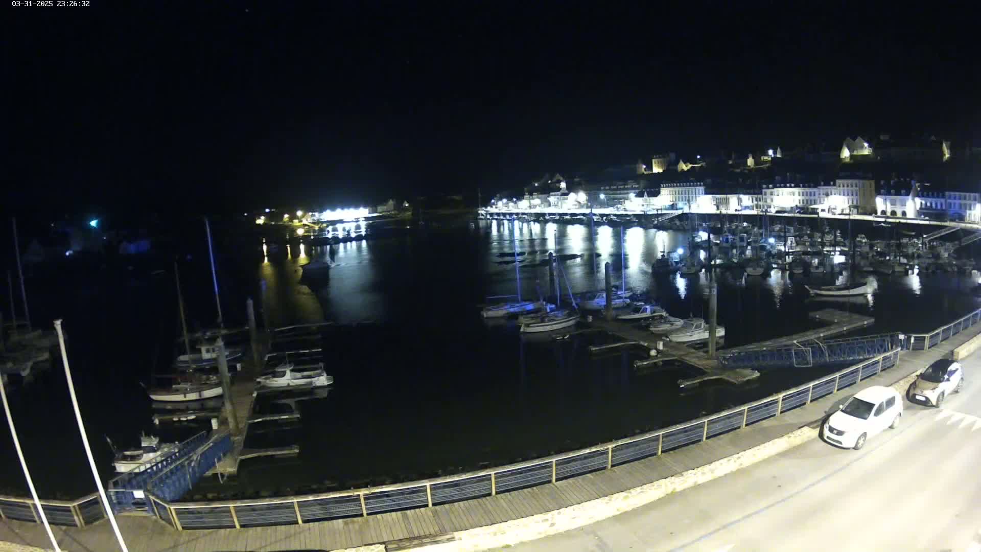A nighttime view of a harbor filled with boats docked along a waterfront, with a town illuminated behind it under a clear, dark sky.
