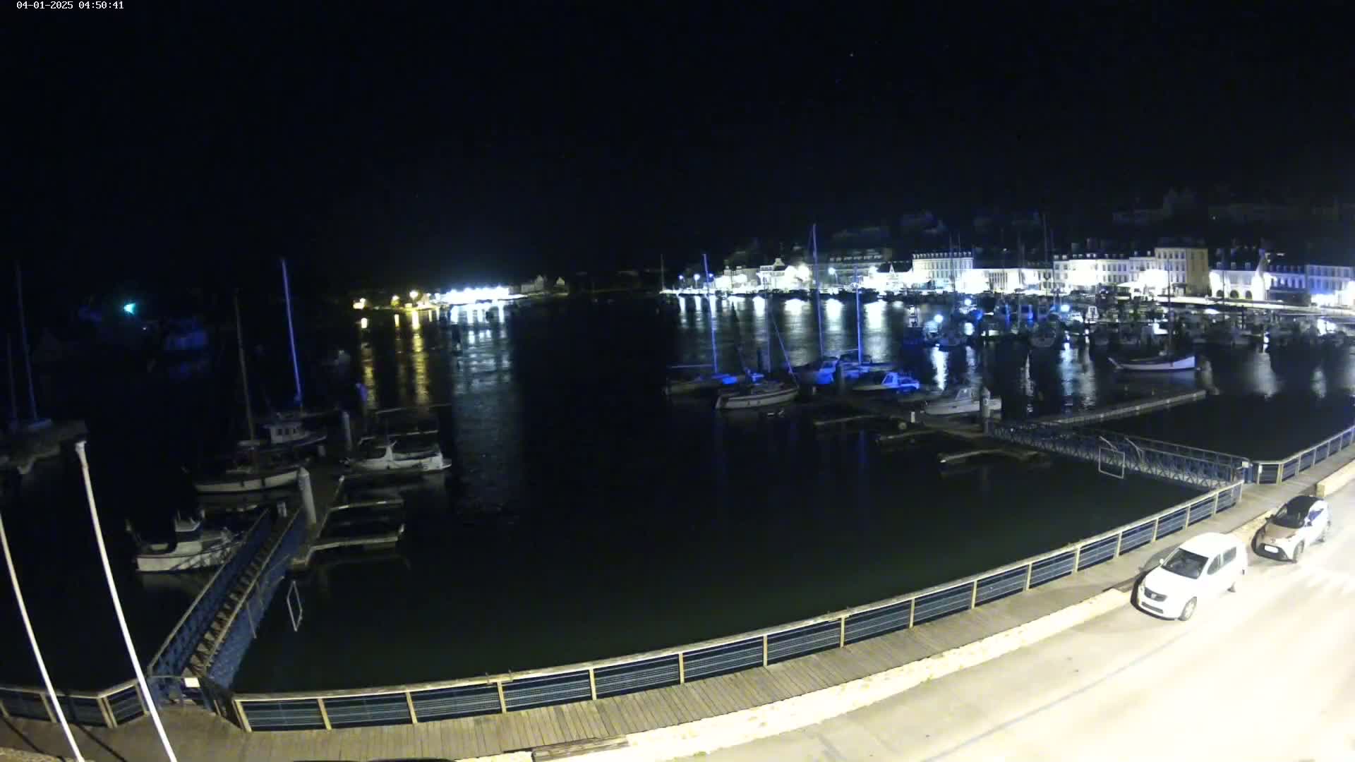 A nighttime view of a harbor filled with boats, docked alongside a waterfront with two cars parked nearby.