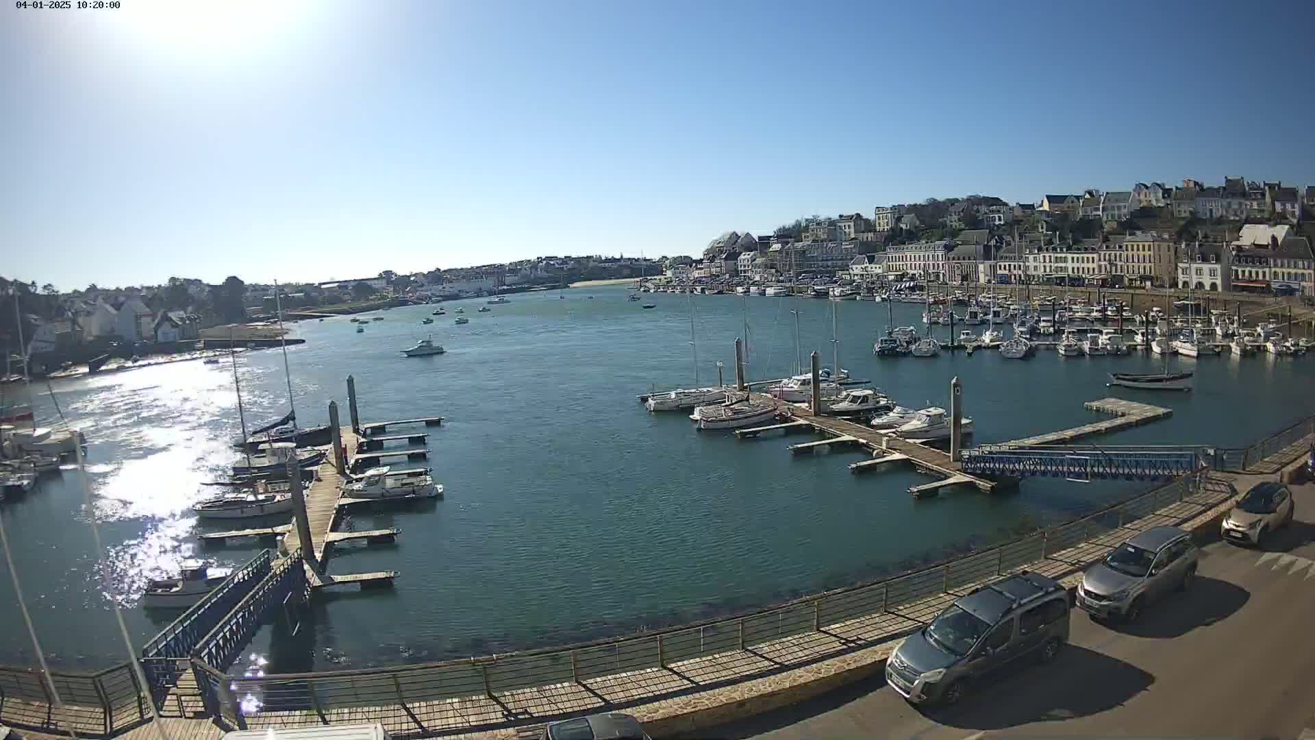 A harbor filled with numerous boats and yachts is situated below a town of white buildings under a bright, sunny sky.