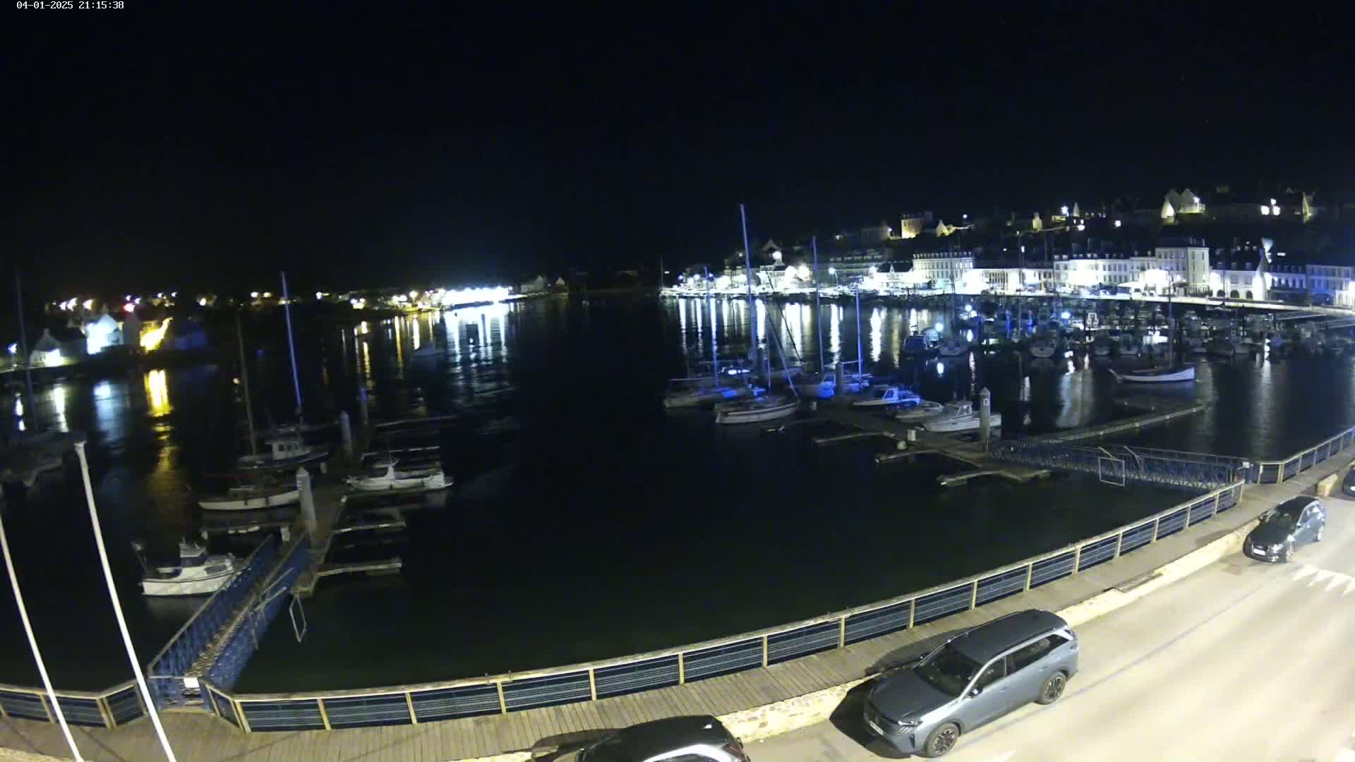 A nighttime view of a harbor filled with boats moored at docks, with a town's lights reflected in the calm water.