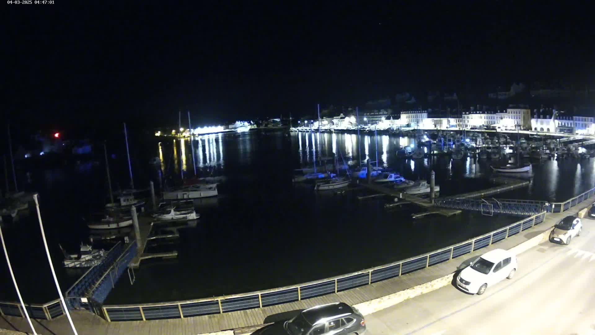 At night, numerous boats are docked in a harbor beside a town with illuminated buildings, under a clear, dark sky.