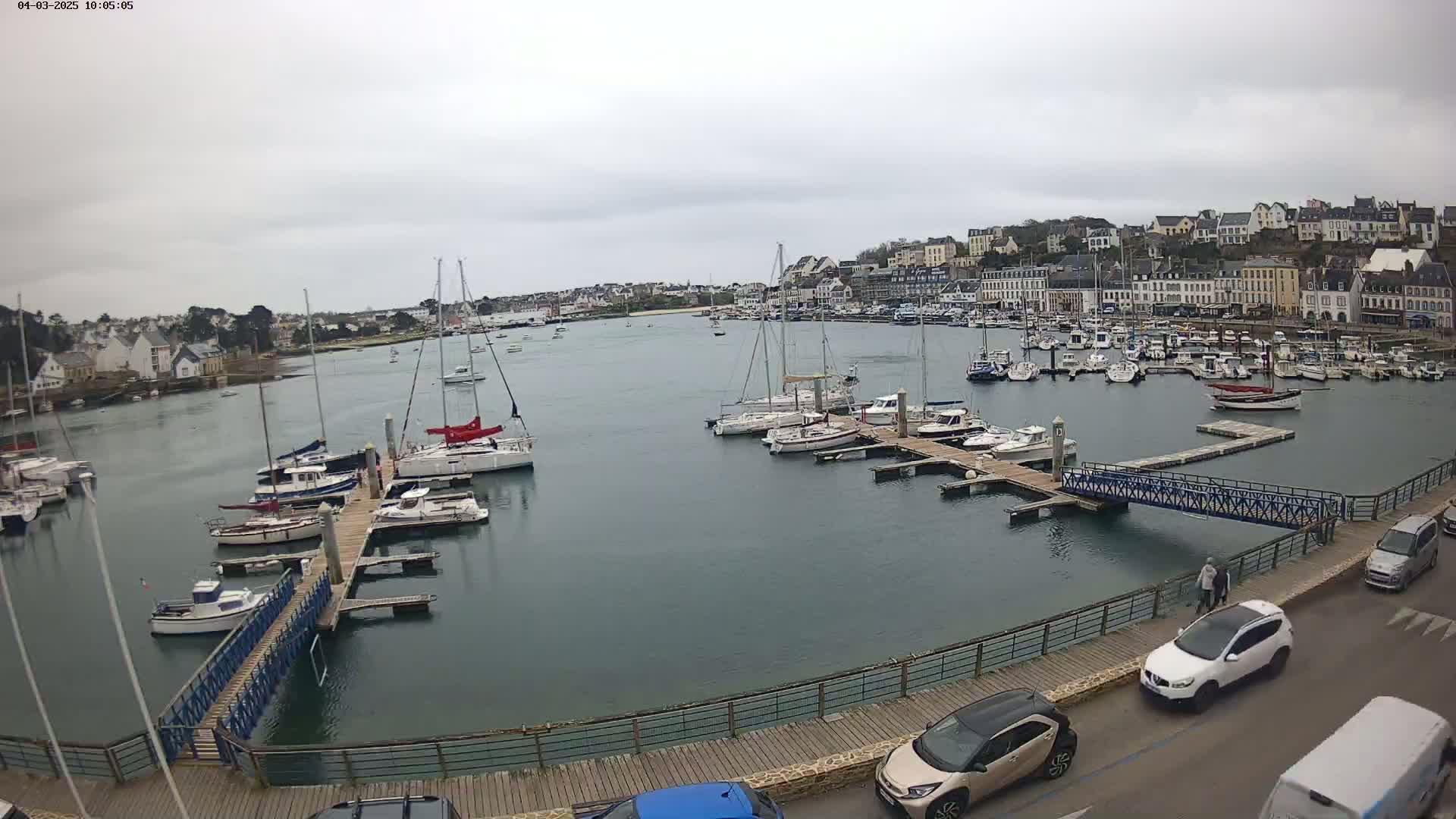 A harbor filled with sailboats and motorboats under an overcast sky, with a town visible in the background and cars parked along a nearby roadway.