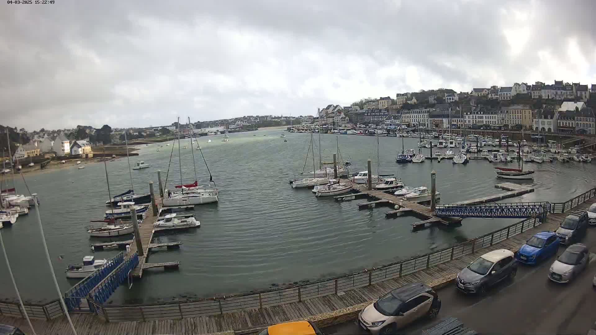 A harbor filled with sailboats and motorboats under an overcast sky, with a town visible in the background and cars parked along a boardwalk.