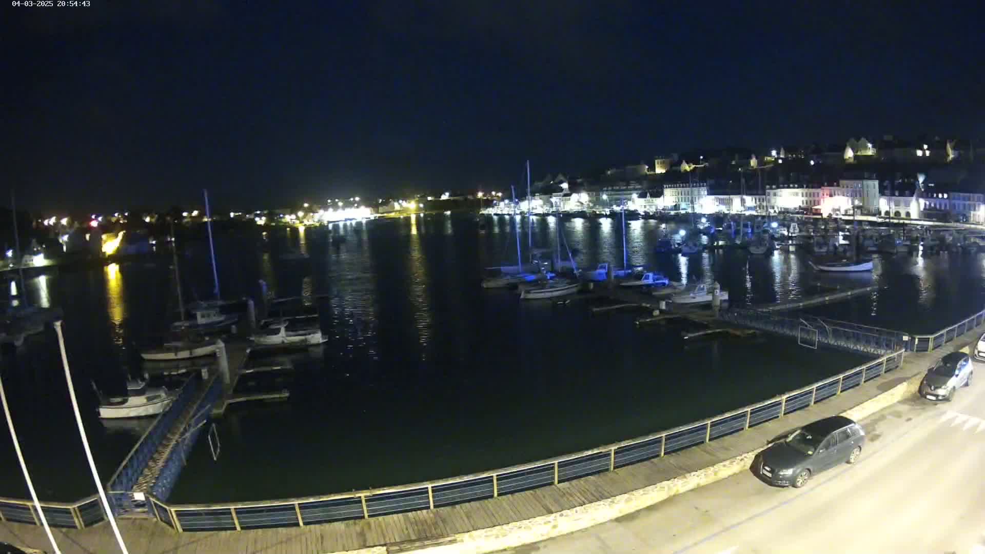 A nighttime view of a harbor filled with boats, docked along a waterfront with a boardwalk and a few parked cars.