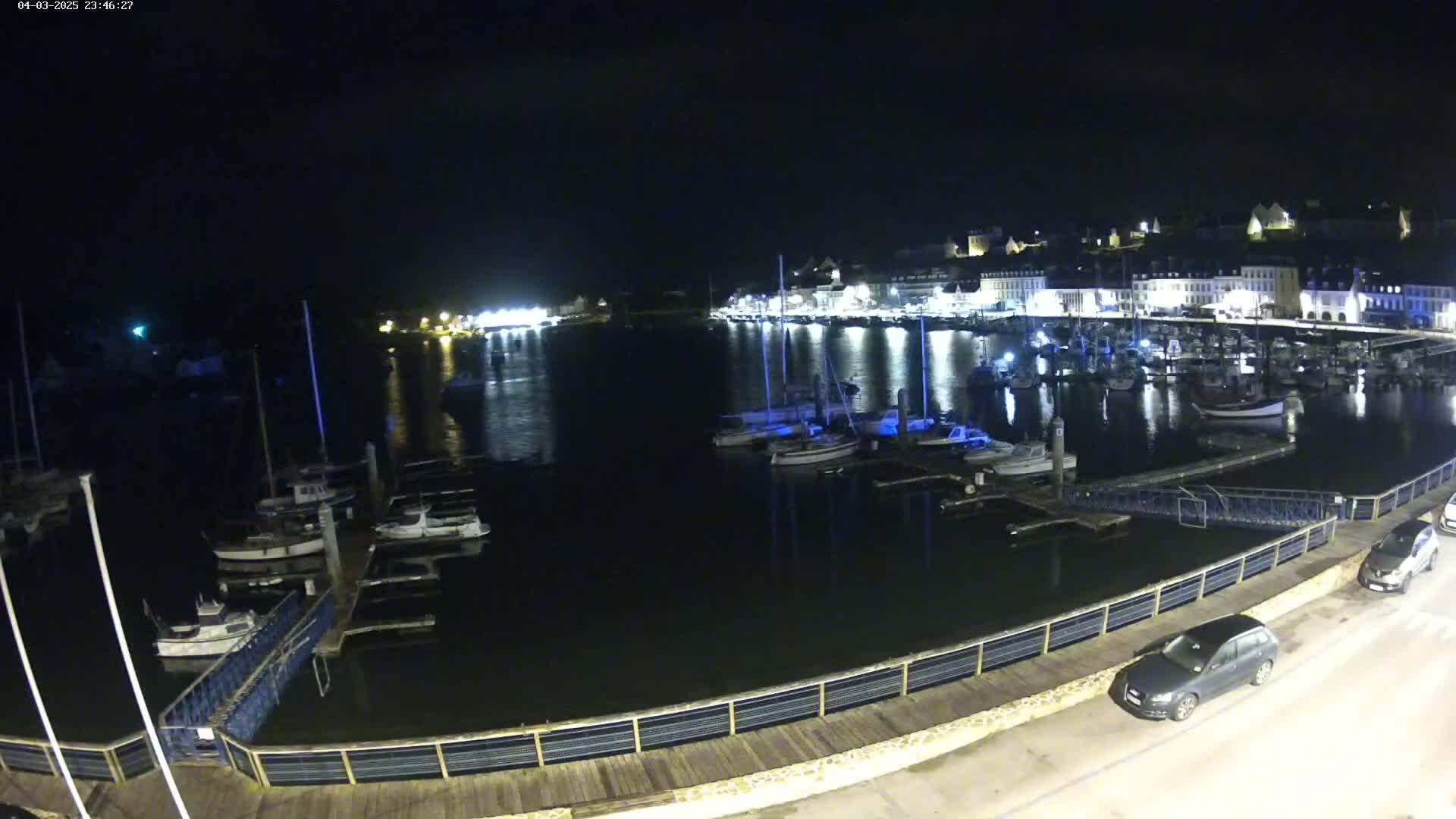 A nighttime view of a harbor filled with docked boats, a waterfront promenade with cars parked alongside it, and a town's lights reflecting on the dark water.