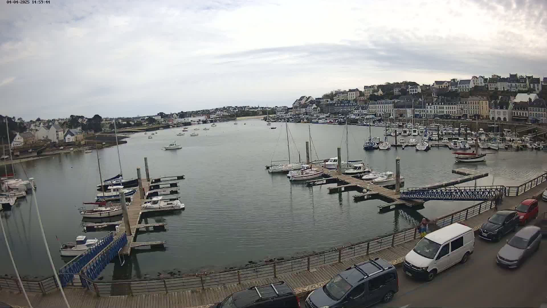 A harbor filled with numerous boats under an overcast sky, with a town in the background and cars parked along a nearby boardwalk.
