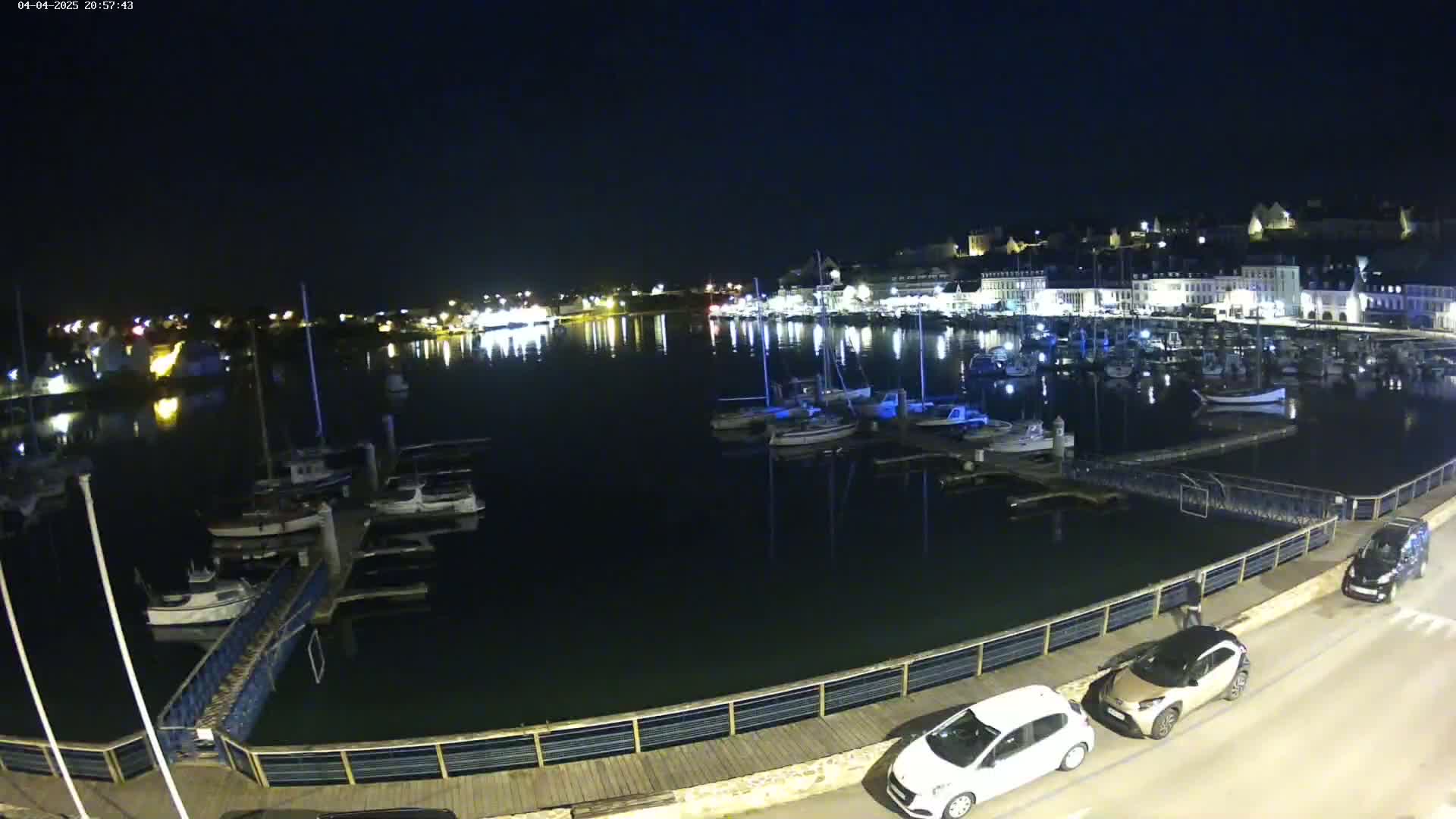 A nighttime view of a harbor filled with boats, a town illuminated with lights in the background, and cars parked along a waterfront walkway.