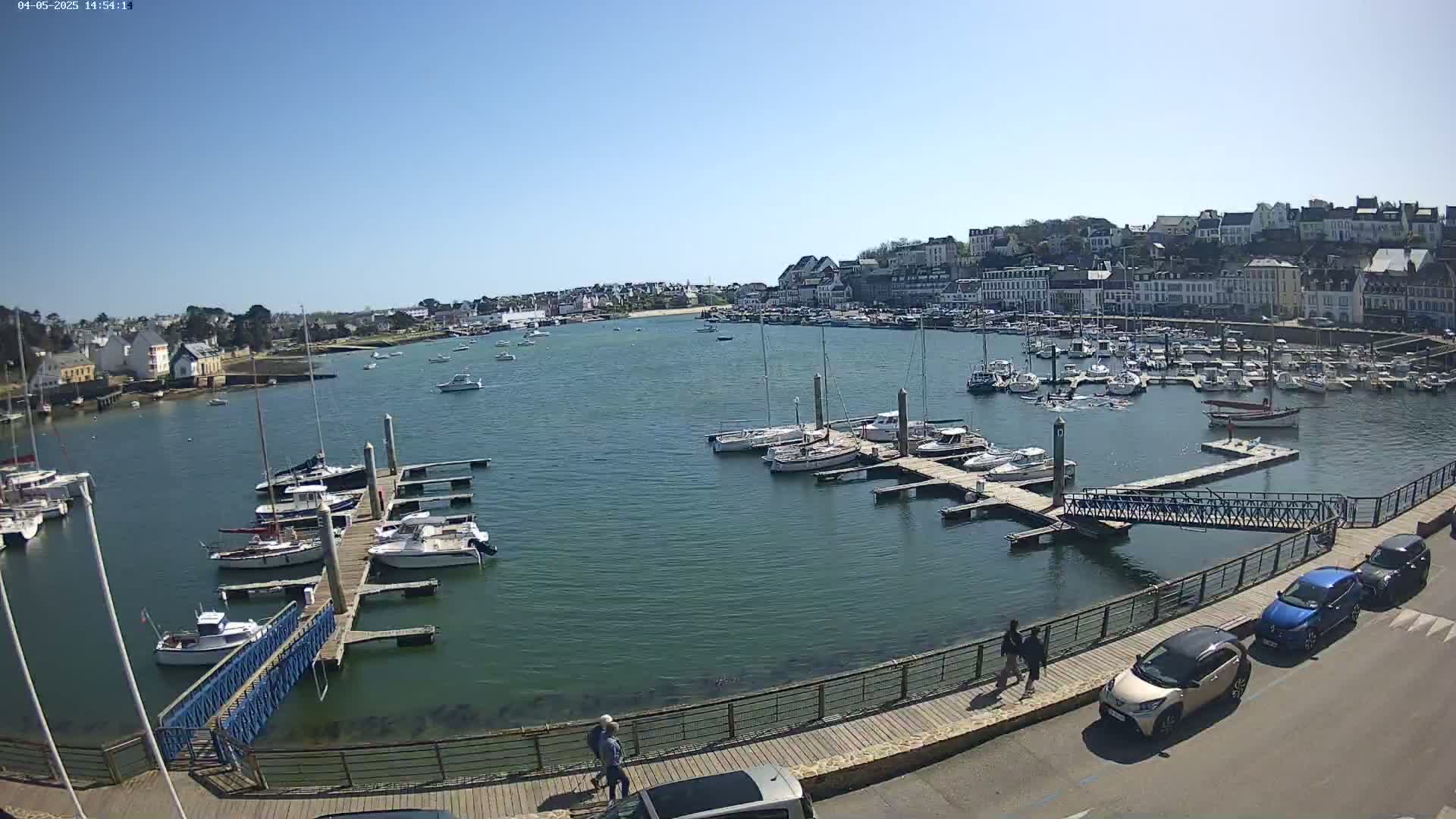 A harbor filled with numerous boats and yachts on a sunny day, with a town's buildings visible in the background.