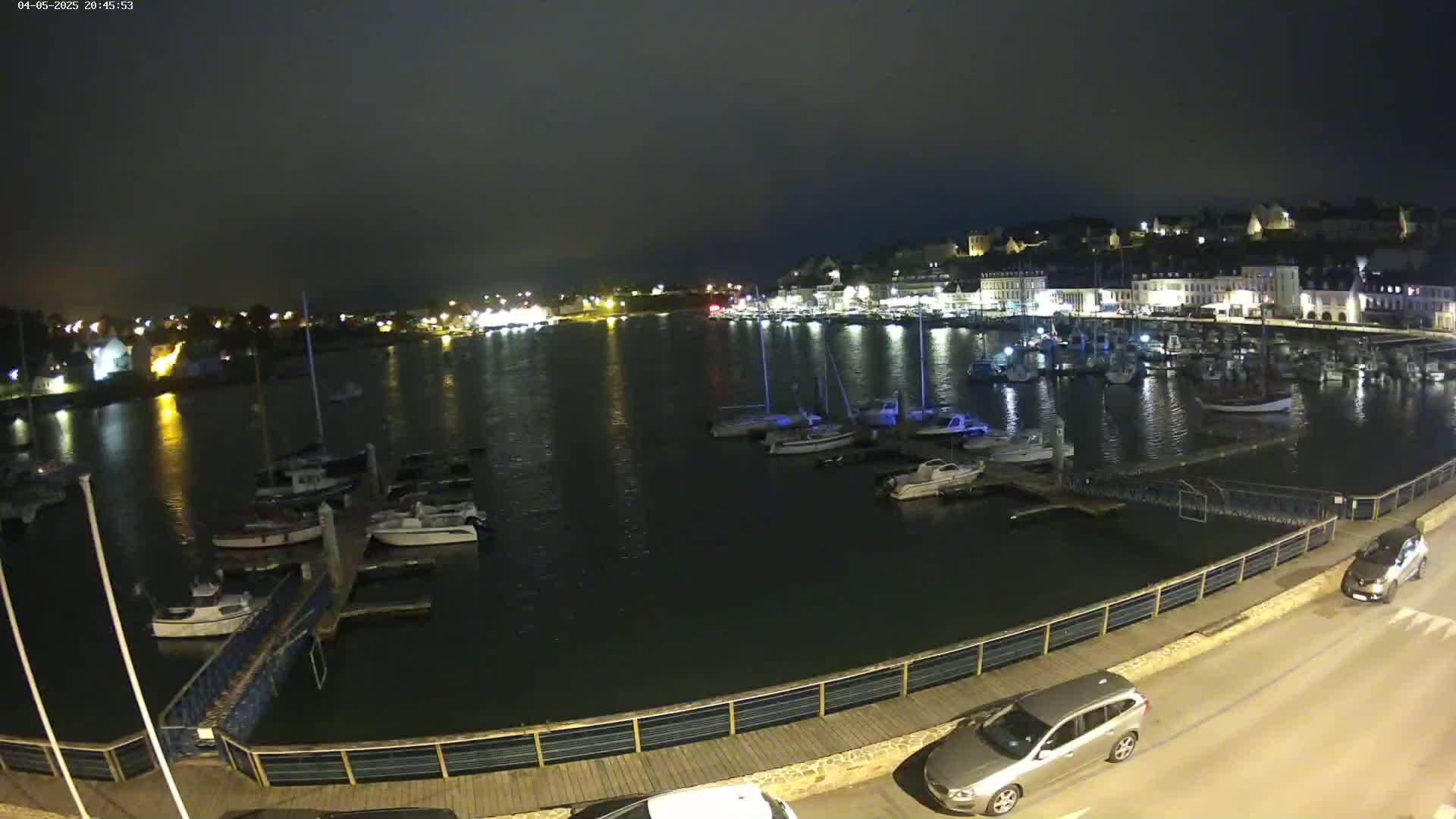 A nighttime view of a harbor filled with boats, a town with illuminated buildings in the background, and a roadway with cars parked alongside a waterfront walkway.