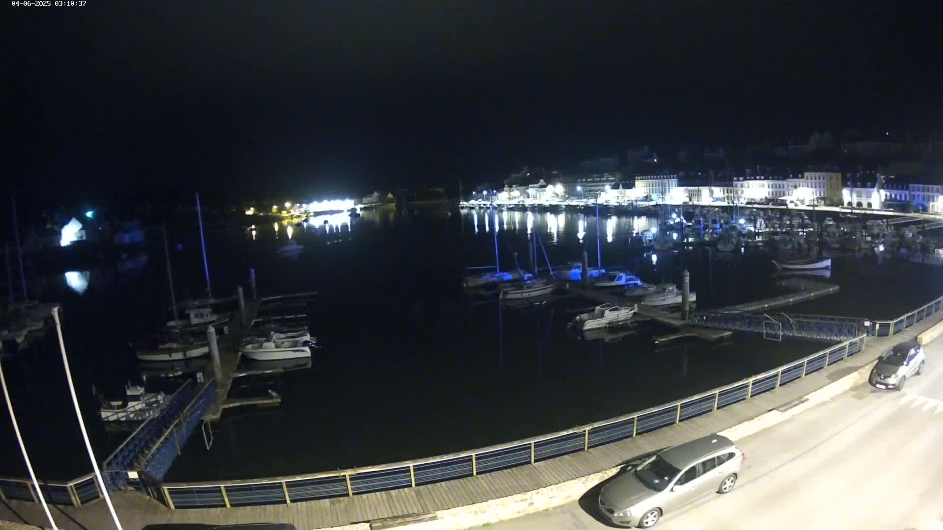 At night, a harbor filled with boats is illuminated, with a town's lights reflecting on the calm water.