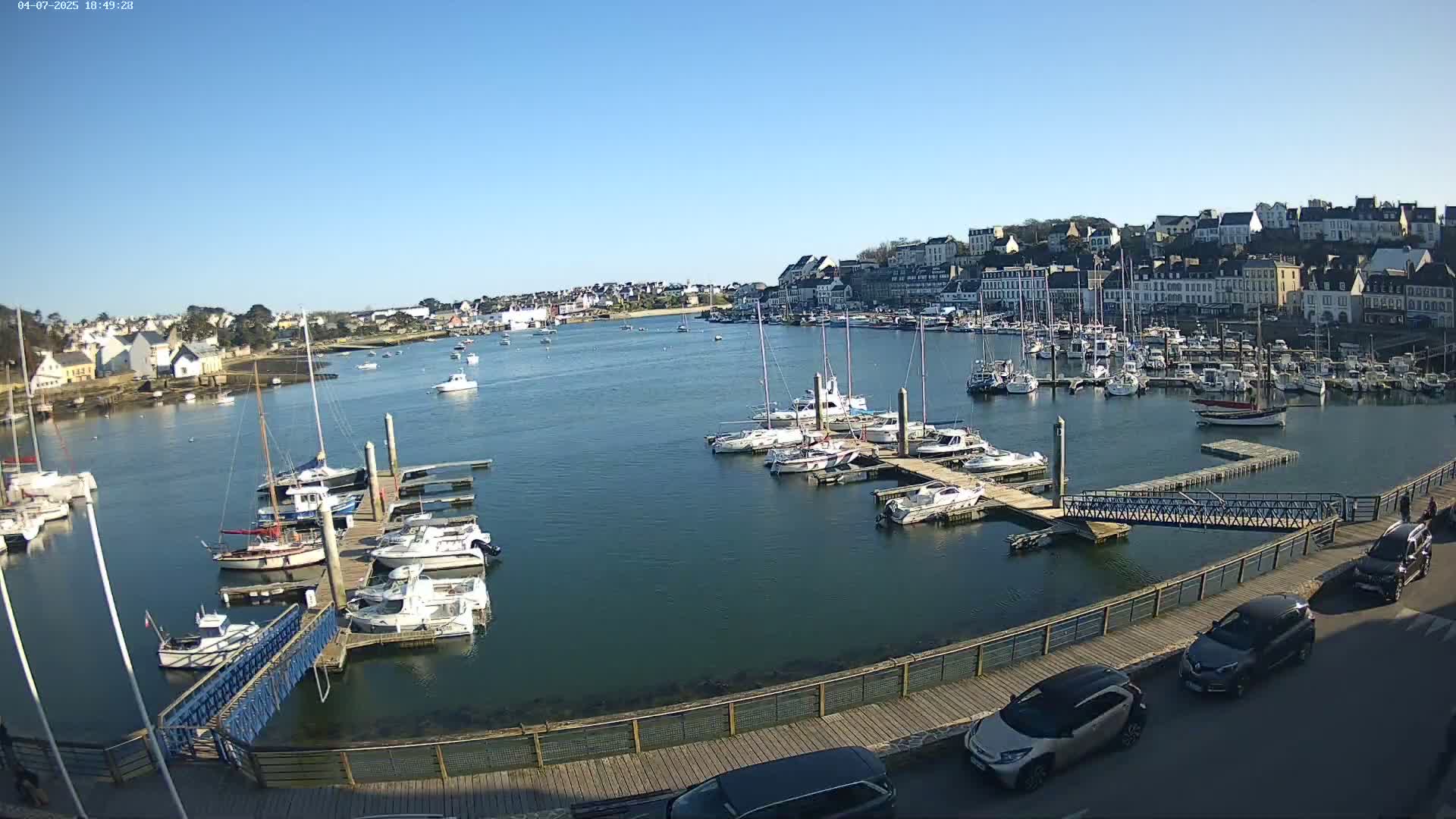 A harbor filled with numerous boats and yachts under a clear, sunny sky, with a town visible in the background.