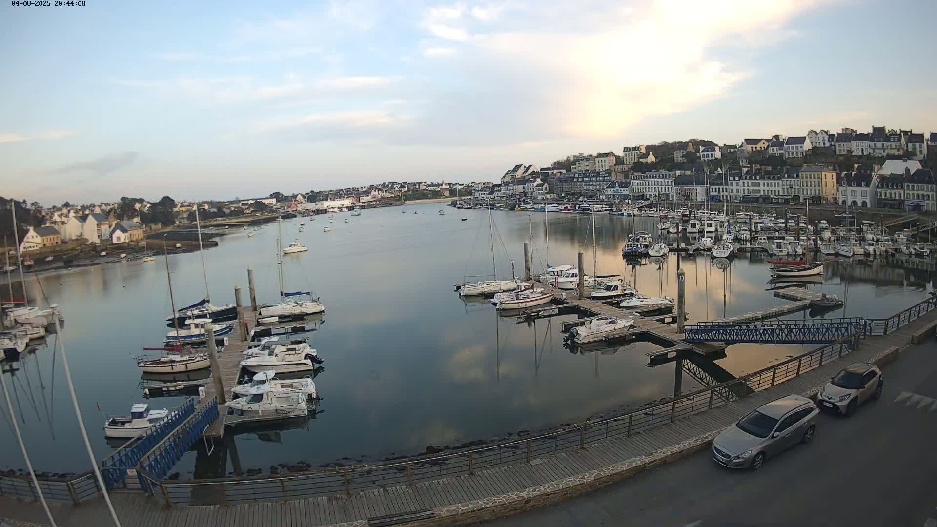A calm harbor filled with numerous sailboats docked at piers, under a twilight sky, with a town visible in the background.