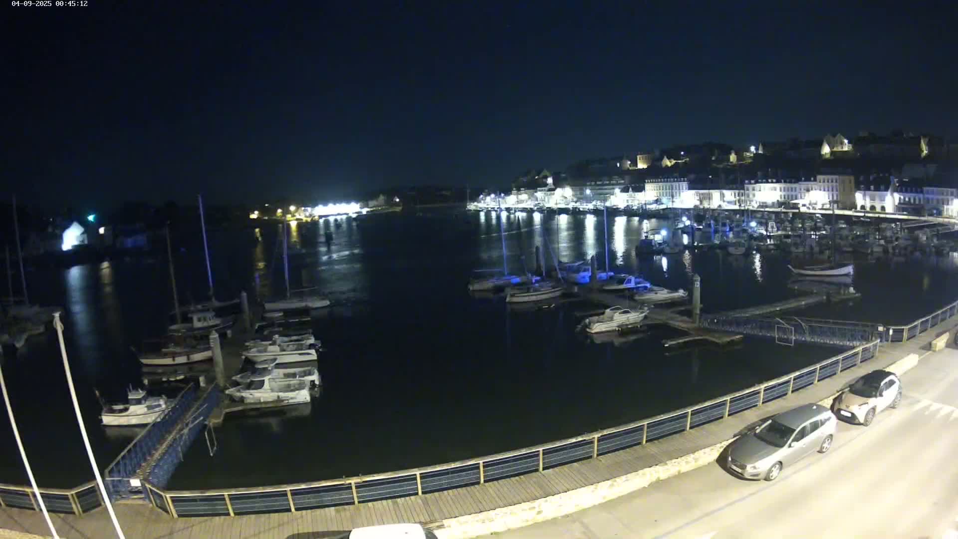 A nighttime view of a harbor filled with boats, docked along a waterfront with a boardwalk and two cars parked nearby under a clear, dark sky.