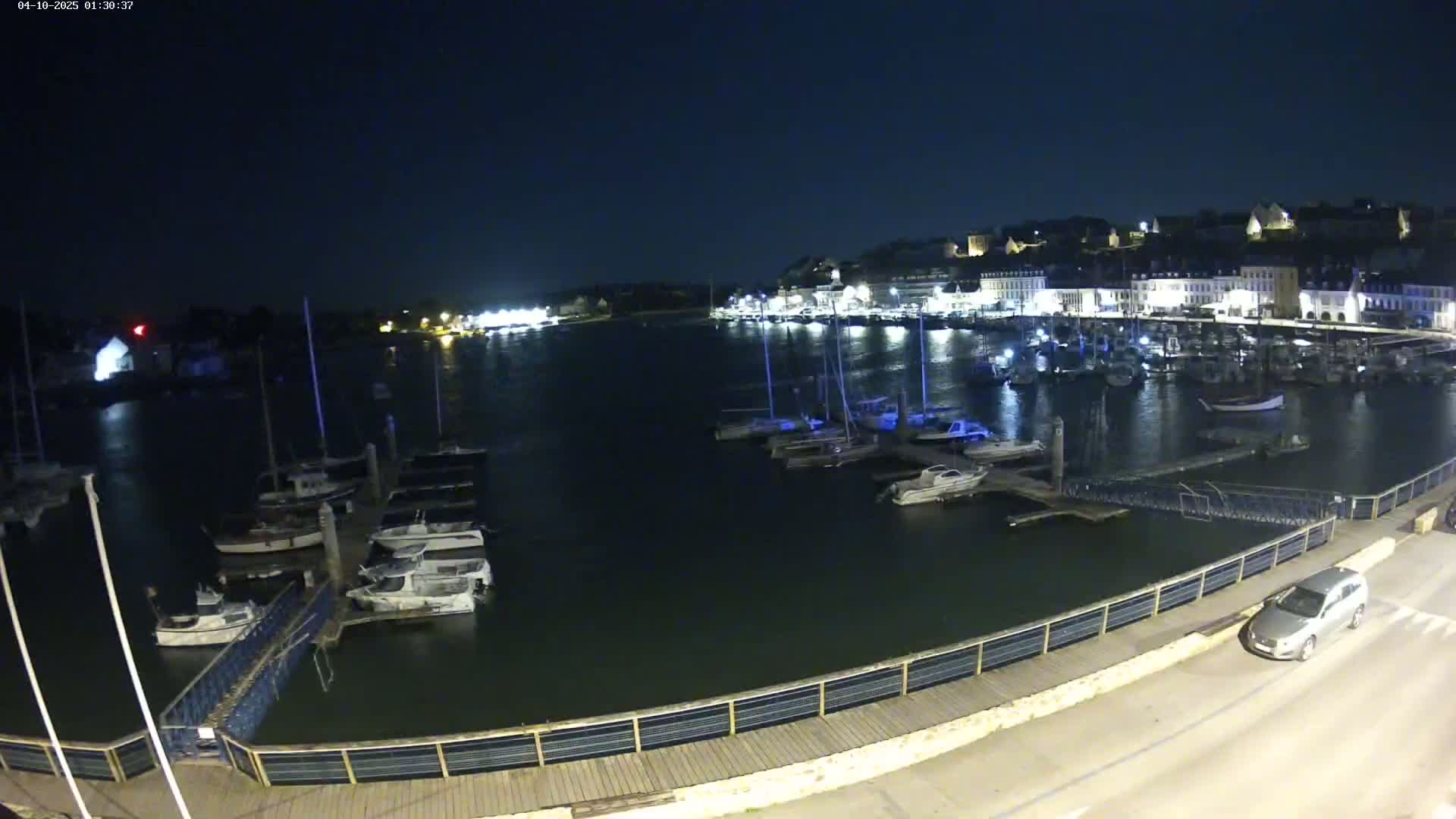 A nighttime view of a harbor filled with boats docked along a waterfront, with a town illuminated in the background under a clear, dark sky.