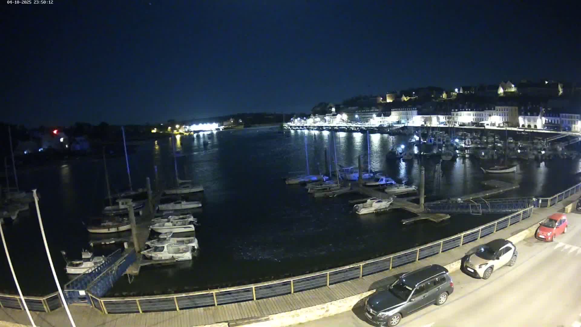 A nighttime view of a harbor filled with boats, docked along a waterfront with cars parked nearby under a clear, dark sky.