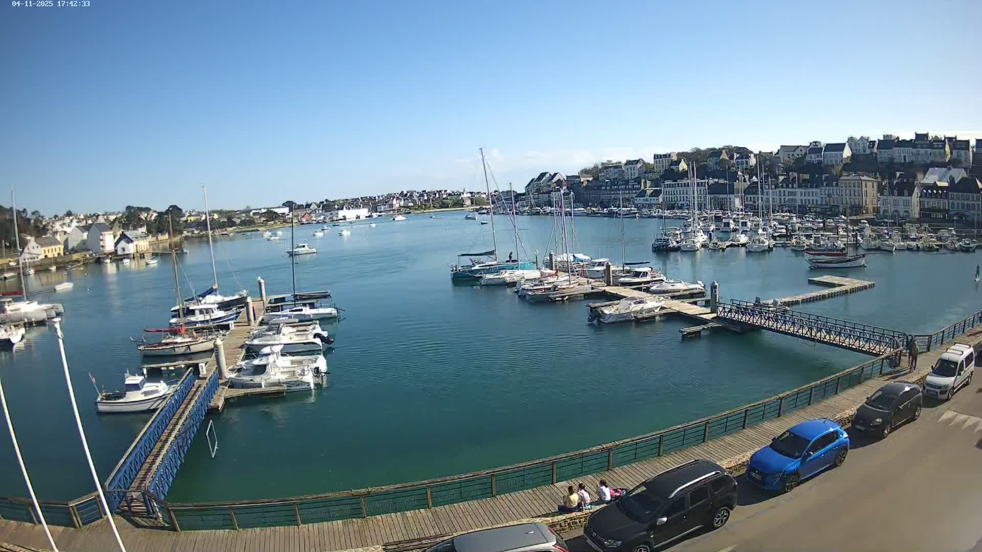 A harbor filled with sailboats and motorboats on a sunny day, with a town's buildings visible in the background.