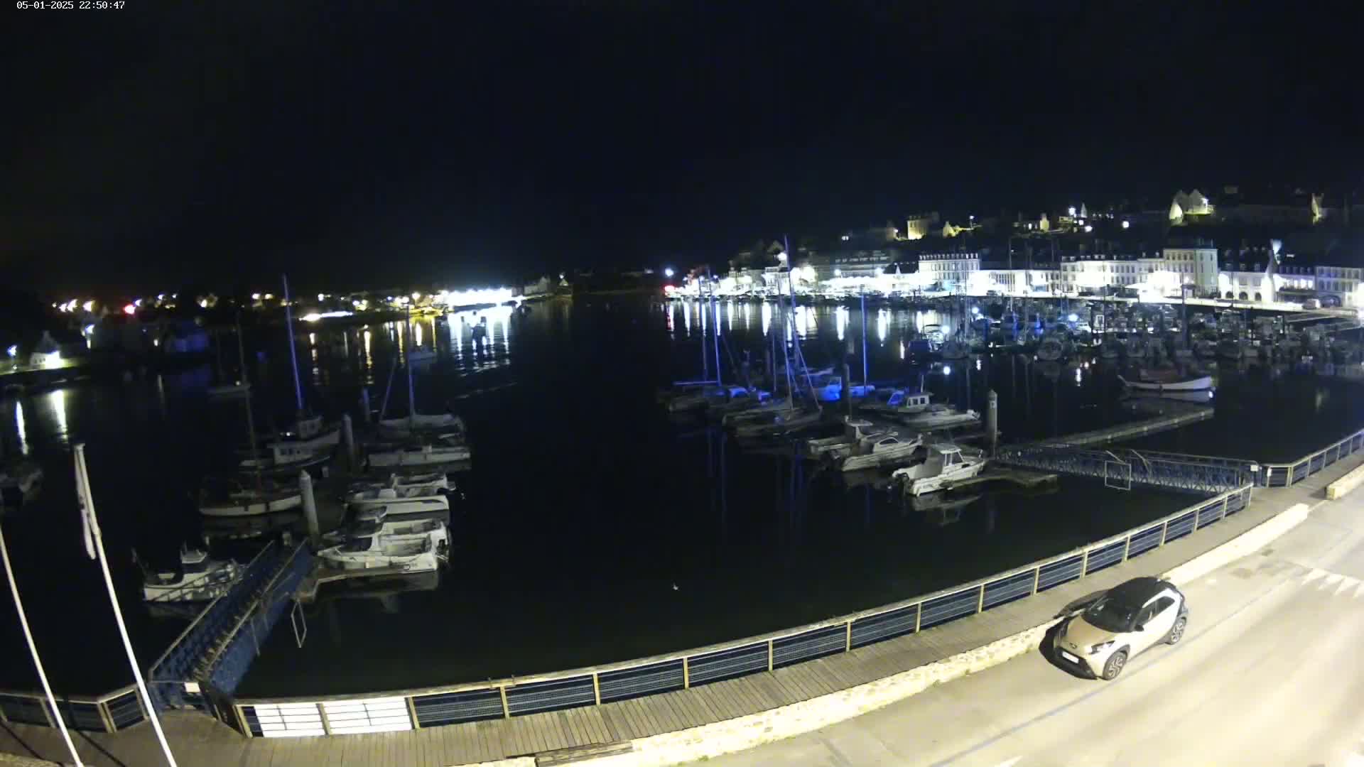 A nighttime view of a harbor filled with boats, with a town's lights reflecting on the dark water, and a single car parked nearby.