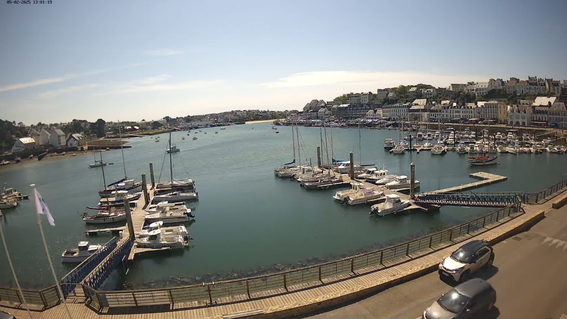 A sunny harbor filled with sailboats and motorboats docked at piers, with a town and hills visible in the background.