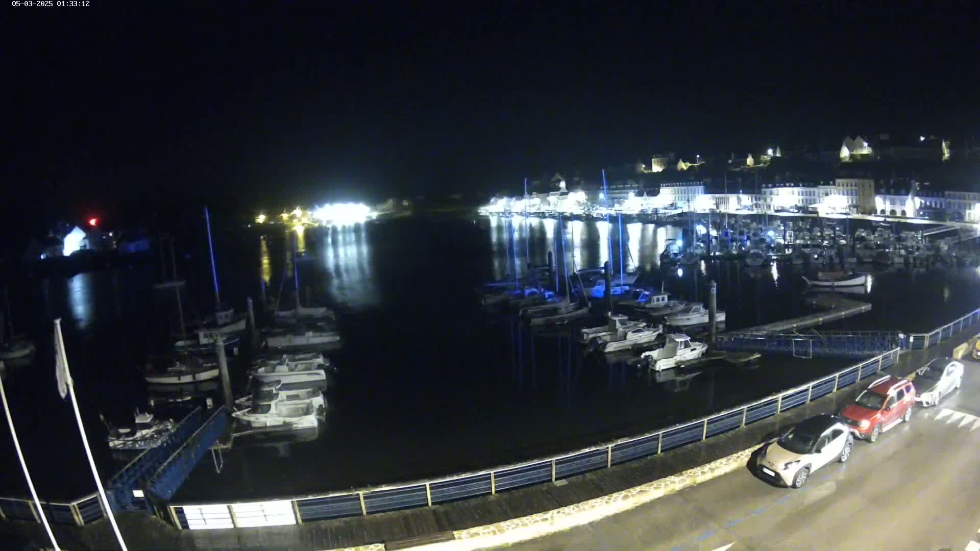 A nighttime view of a harbor filled with boats, next to a town with buildings illuminated, under a dark sky.