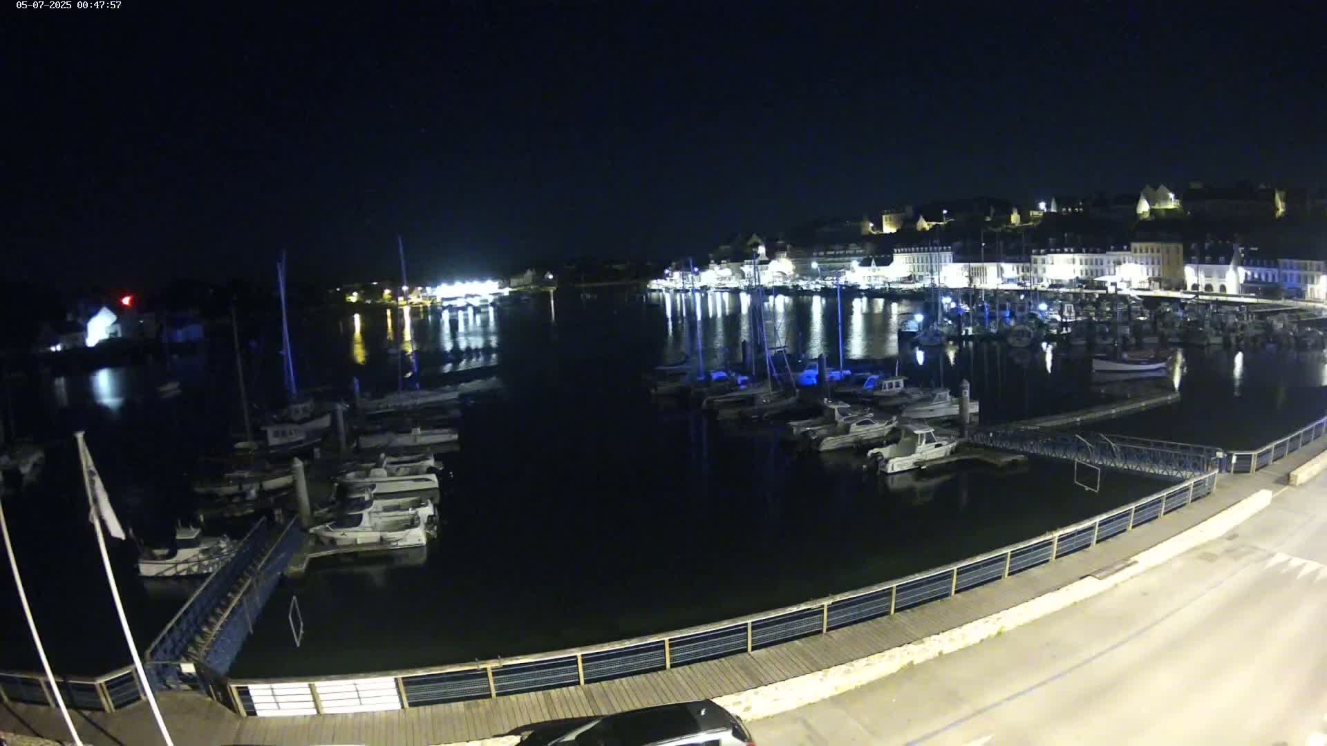 A nighttime view of a harbor filled with boats, reflecting the lights of a town across the water under a clear, dark sky.