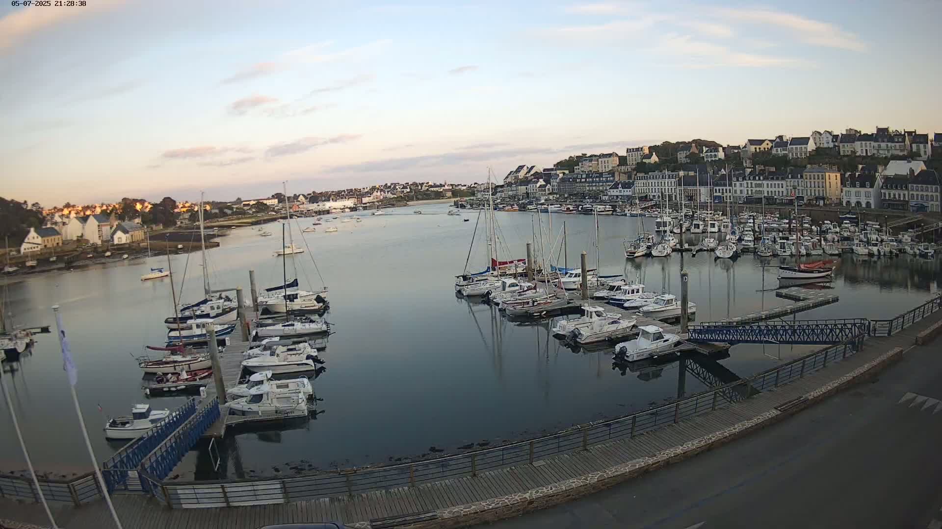 A calm harbor filled with numerous sailboats and motorboats at sunset, with a town of white buildings visible in the background.