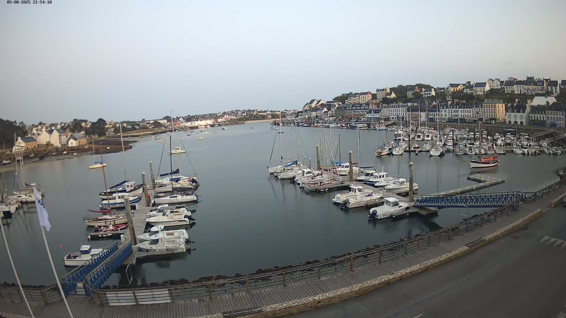 A calm harbor filled with many sailboats and motorboats is nestled next to a town under a clear, pale sky.