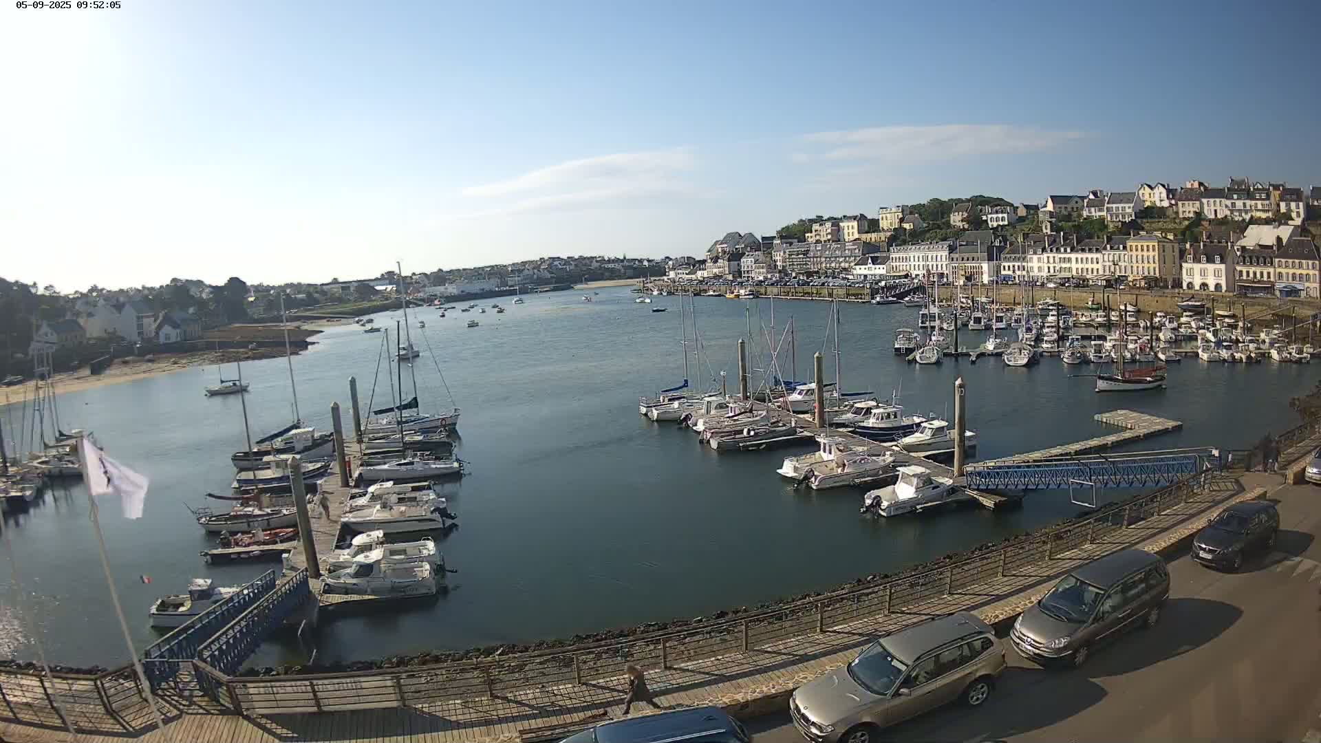 A harbor filled with sailboats and motorboats on a sunny day, with a town and parked cars visible in the background.