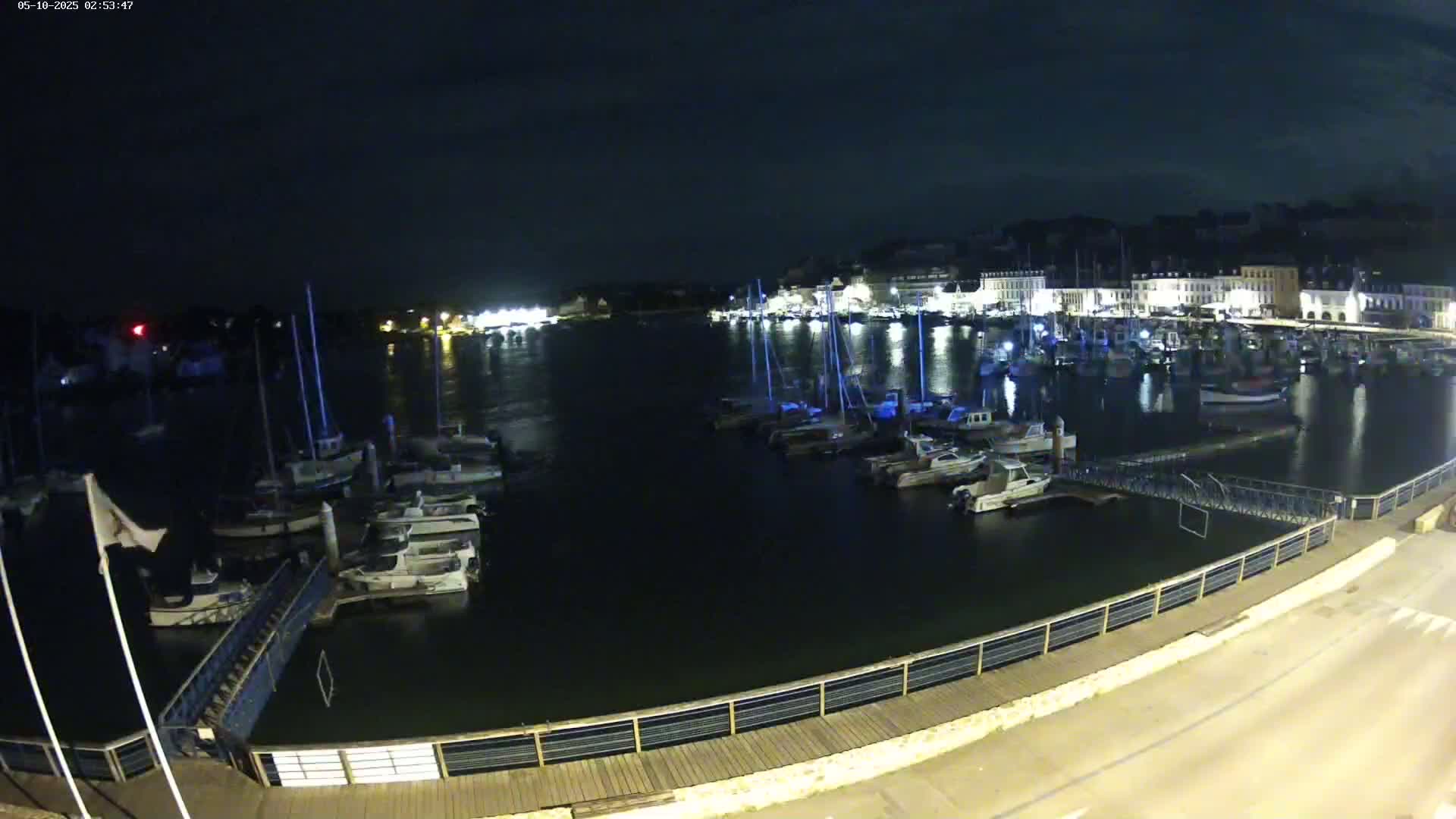 At night, a harbor filled with boats is seen from a waterfront boardwalk under a dark sky.