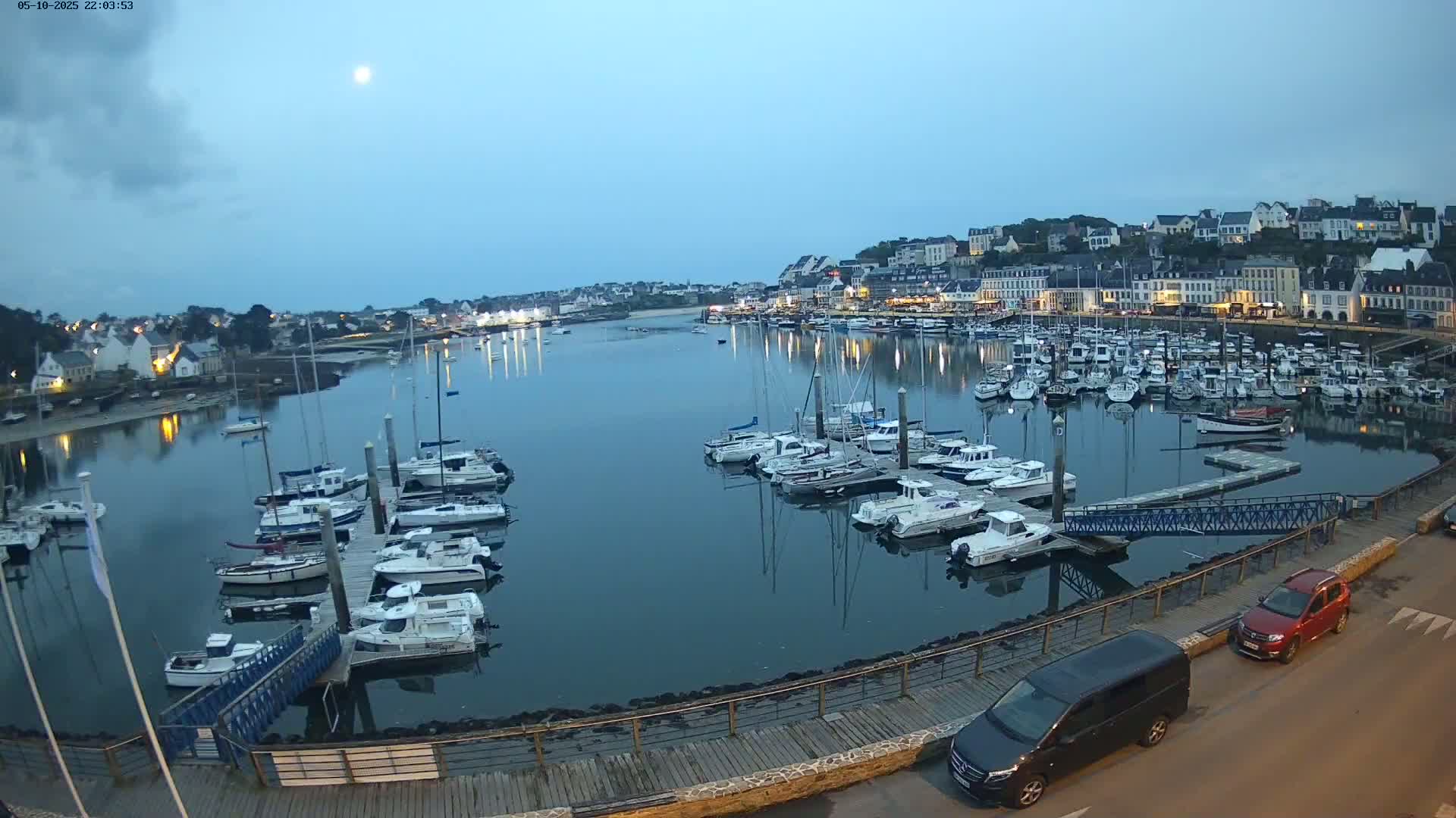 A harbor filled with numerous boats at dusk, under a mostly clear sky with a visible moon, is nestled next to a town with buildings lining the waterfront.