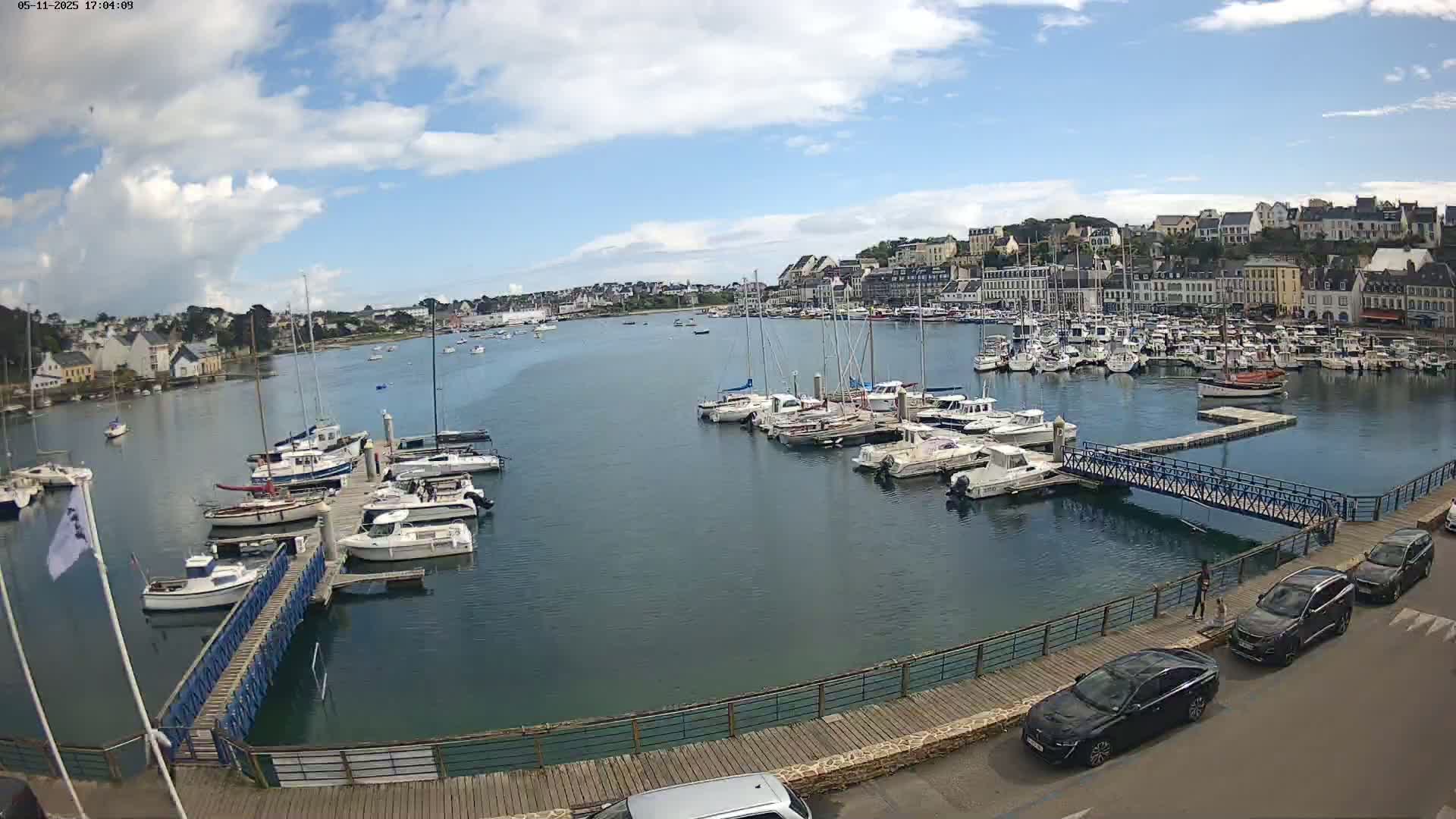 A harbor filled with numerous boats and yachts under a partly cloudy sky is viewed from a waterfront boardwalk with several cars parked nearby.