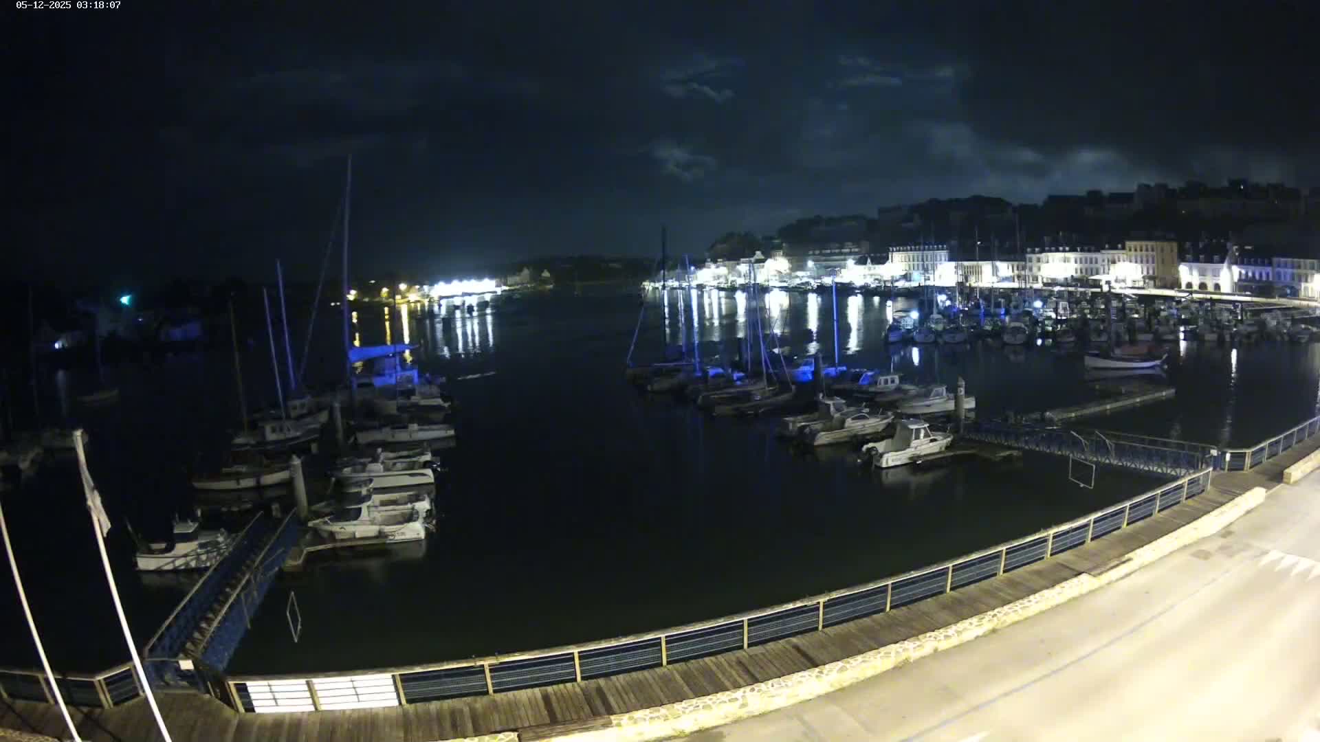 A nighttime view of a harbor filled with boats, illuminated by lights from buildings along the shore under a partly cloudy sky.
