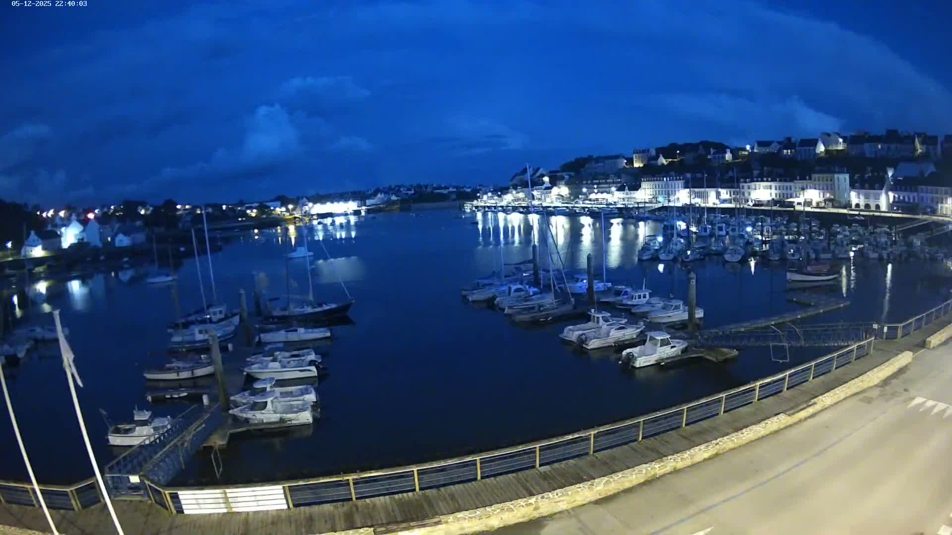 A nighttime view of a harbor filled with boats, under a partly cloudy sky, shows a town illuminated on the far shore.
