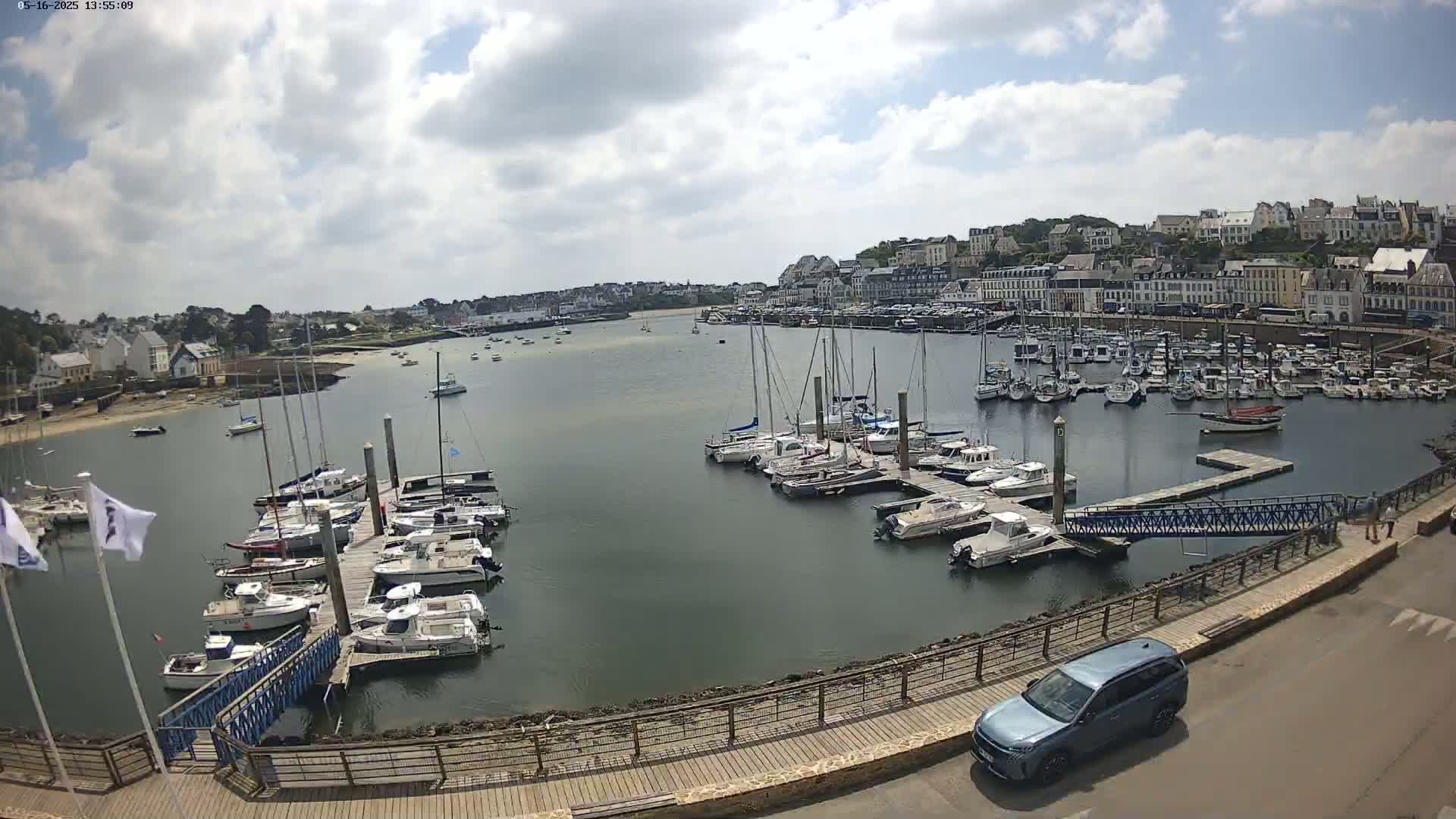A partly cloudy day reveals a harbor filled with numerous sailboats and motorboats docked at piers, with a town and more boats in the background.