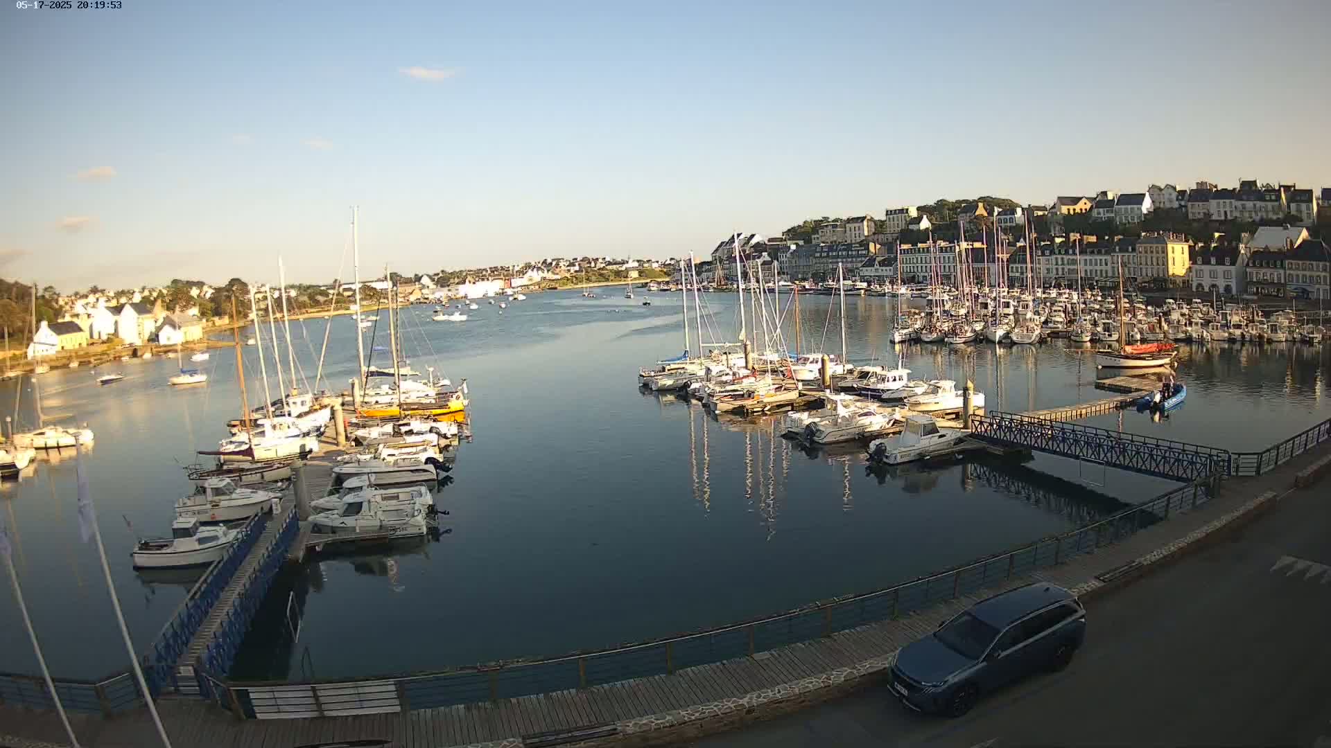 A calm harbor filled with numerous sailboats and motorboats is nestled next to a town under a clear, bright sky.