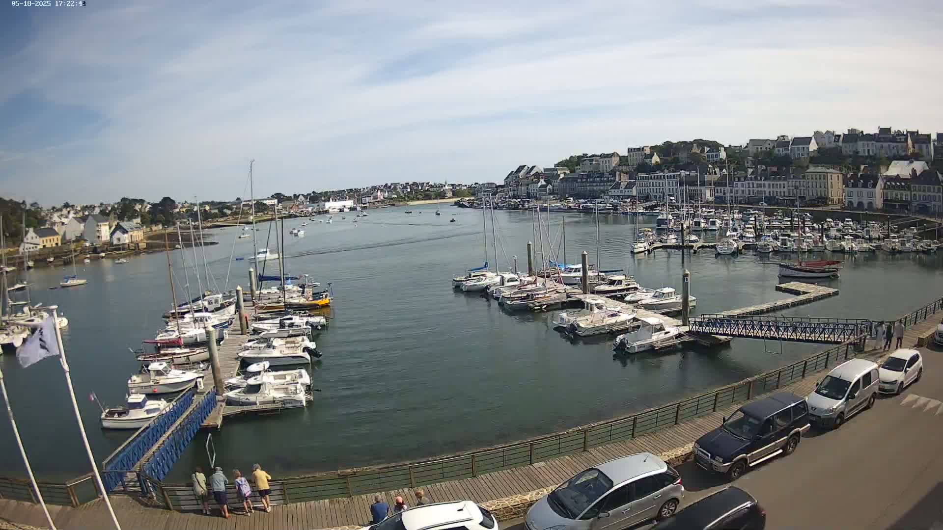 A harbor filled with numerous sailboats and motorboats is seen on a partly sunny day, with several cars parked along a boardwalk overlooking the water and a small group of people standing on the boardwalk.