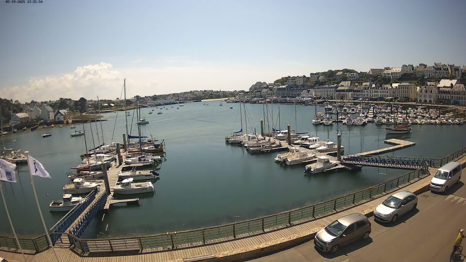 A harbor filled with numerous sailboats and motorboats docked at piers, under a mostly sunny sky, is viewed from an elevated position.