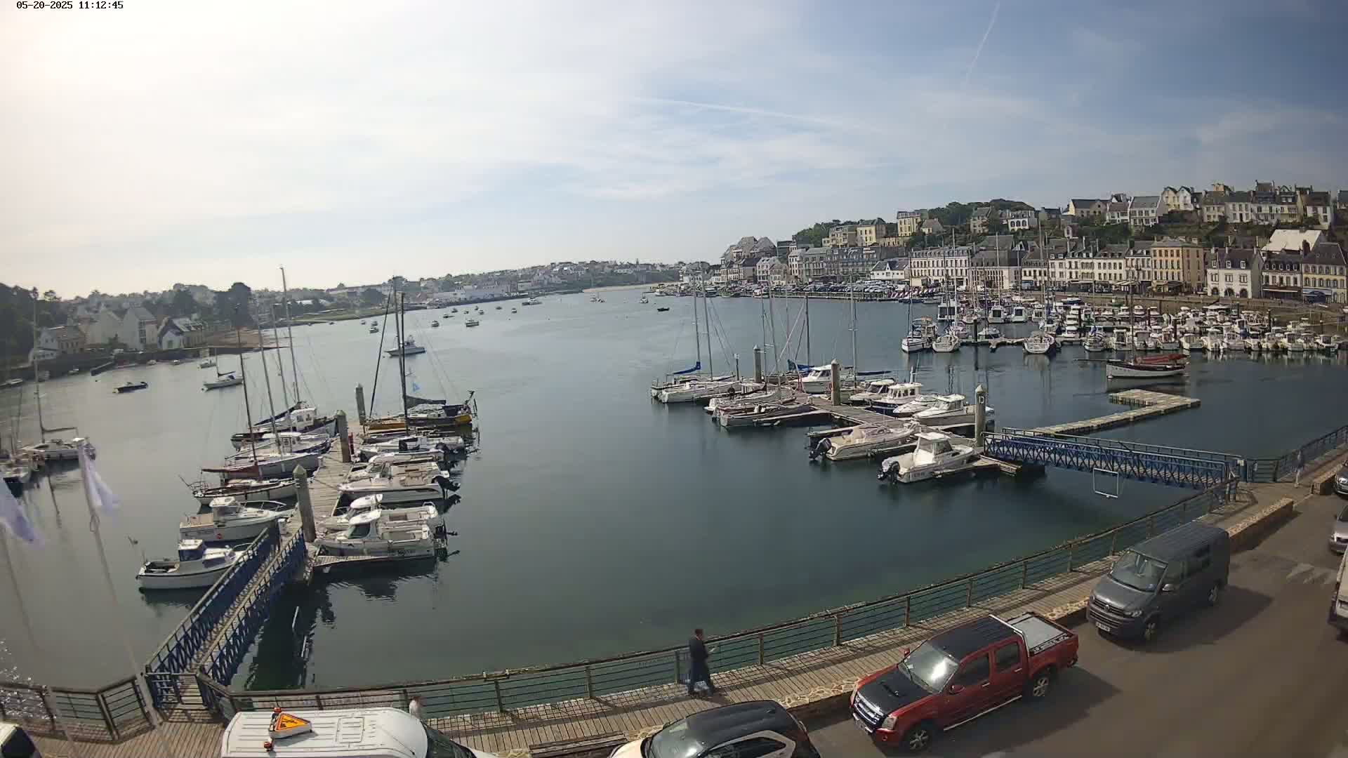 A harbor filled with numerous boats and yachts is seen on a partly sunny day, with a town's buildings visible in the background.