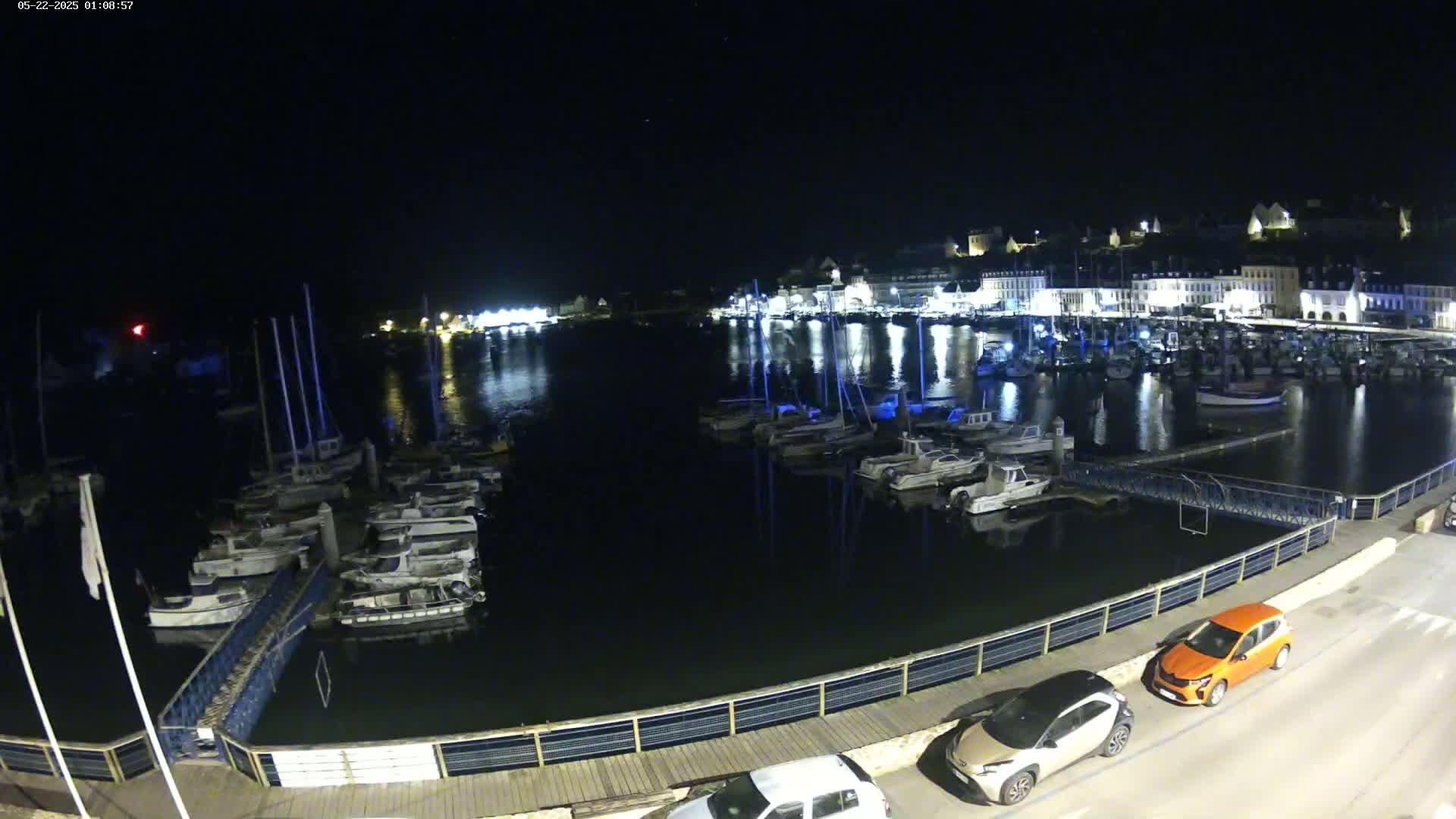 A nighttime harbor scene shows many boats docked along a waterfront, with a town's lights reflecting in the dark water.