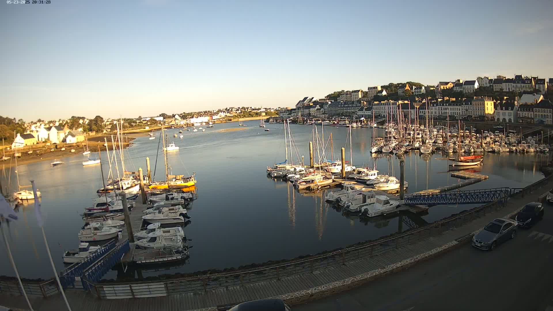 Many sailboats and motorboats are docked in a calm harbor next to a town under a clear, sunny sky.