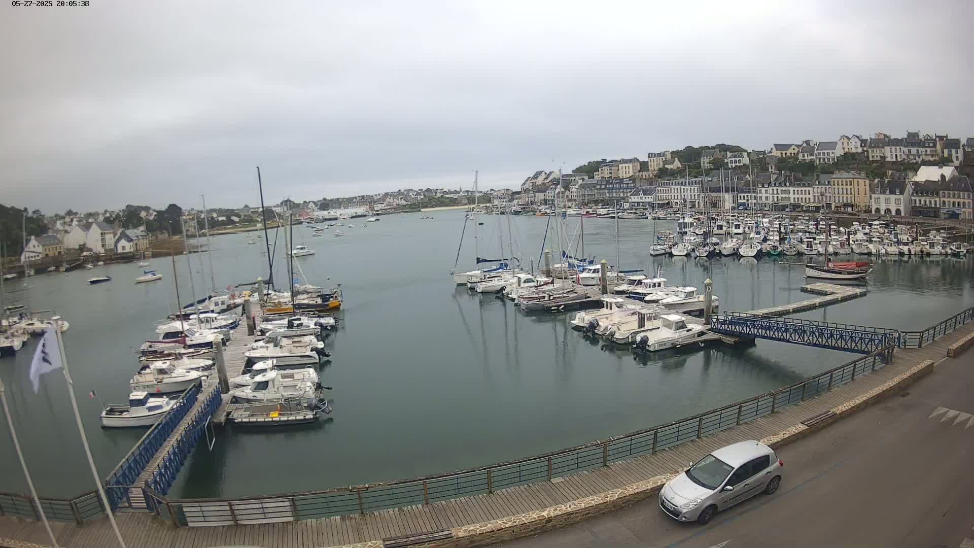 A harbor filled with numerous boats and yachts under a cloudy sky, with a small car parked near a walkway overlooking the water.